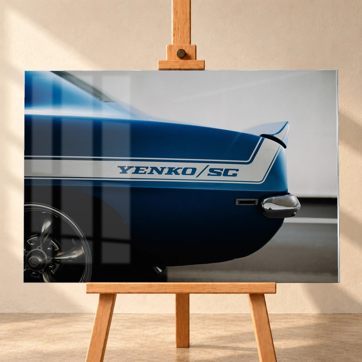 Visualisiertes Acrylglas-Wandbild in frontaler Ansicht auf einer Holzstaffelei vor heller Wand. Fensterspiegelungen liegen über dem Motiv der blauen Heckpartie eines Chevrolet Camaro Yenko mit weißem „YENKO/SC“-Streifen.