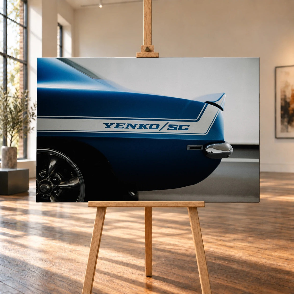 Raumvisualisierung mit querformatigem Leinwandbild auf einer Holzstaffelei in einem hellen Raum mit hohen Fenstern und Holzboden. Das Bild zeigt das blaue Heck eines Chevrolet Camaro Yenko mit weißem „YENKO/SC“-Seitenstreifen.