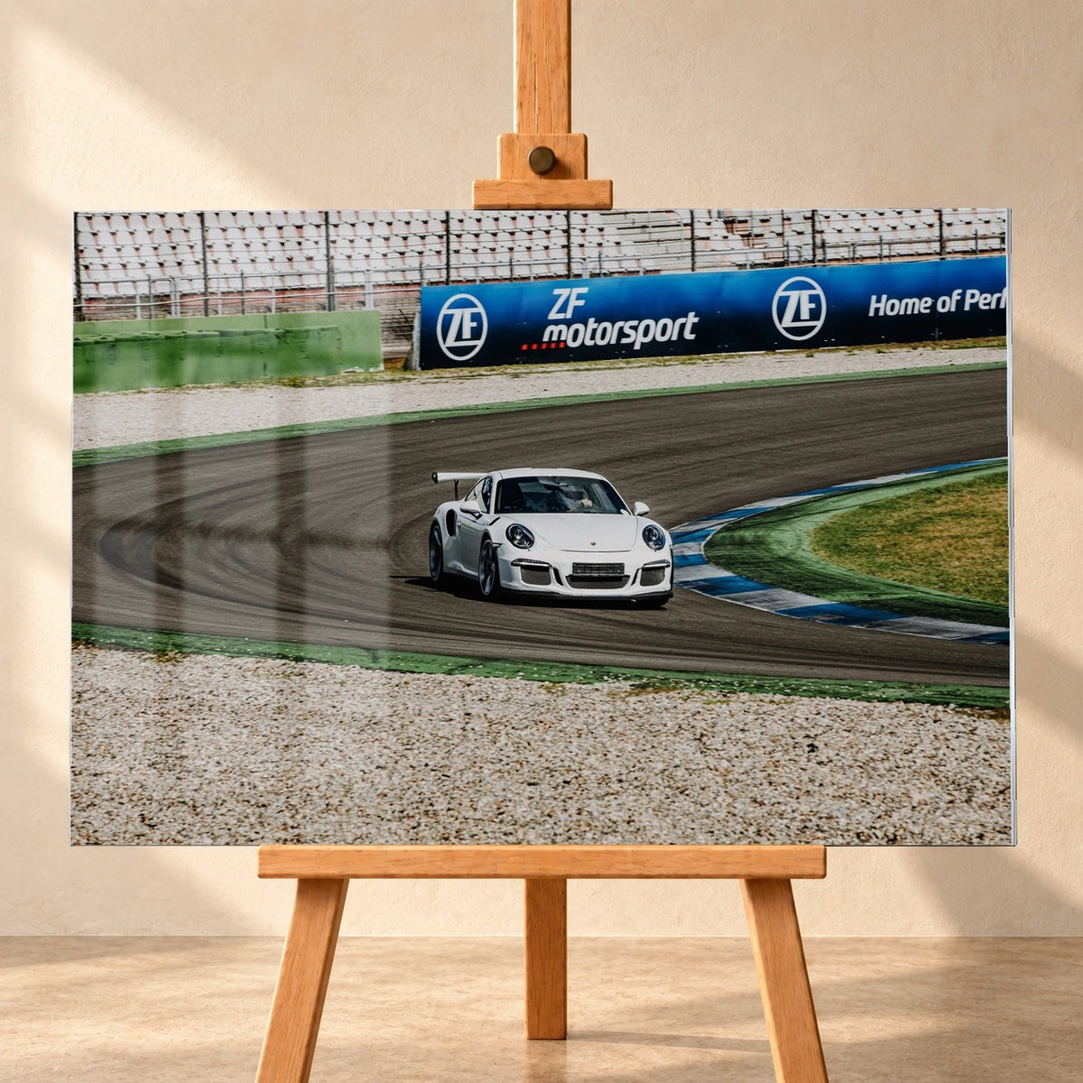 Visualisierung eines Acrylglasbildes von vorn auf einer Holzstaffelei vor heller Wand. Das querformatige Motiv zeigt einen weißen Porsche 911 mit Heckflügel in einer Rennstreckenkurve; links sind Fensterspiegelungen auf der Bildfläche dargestellt.