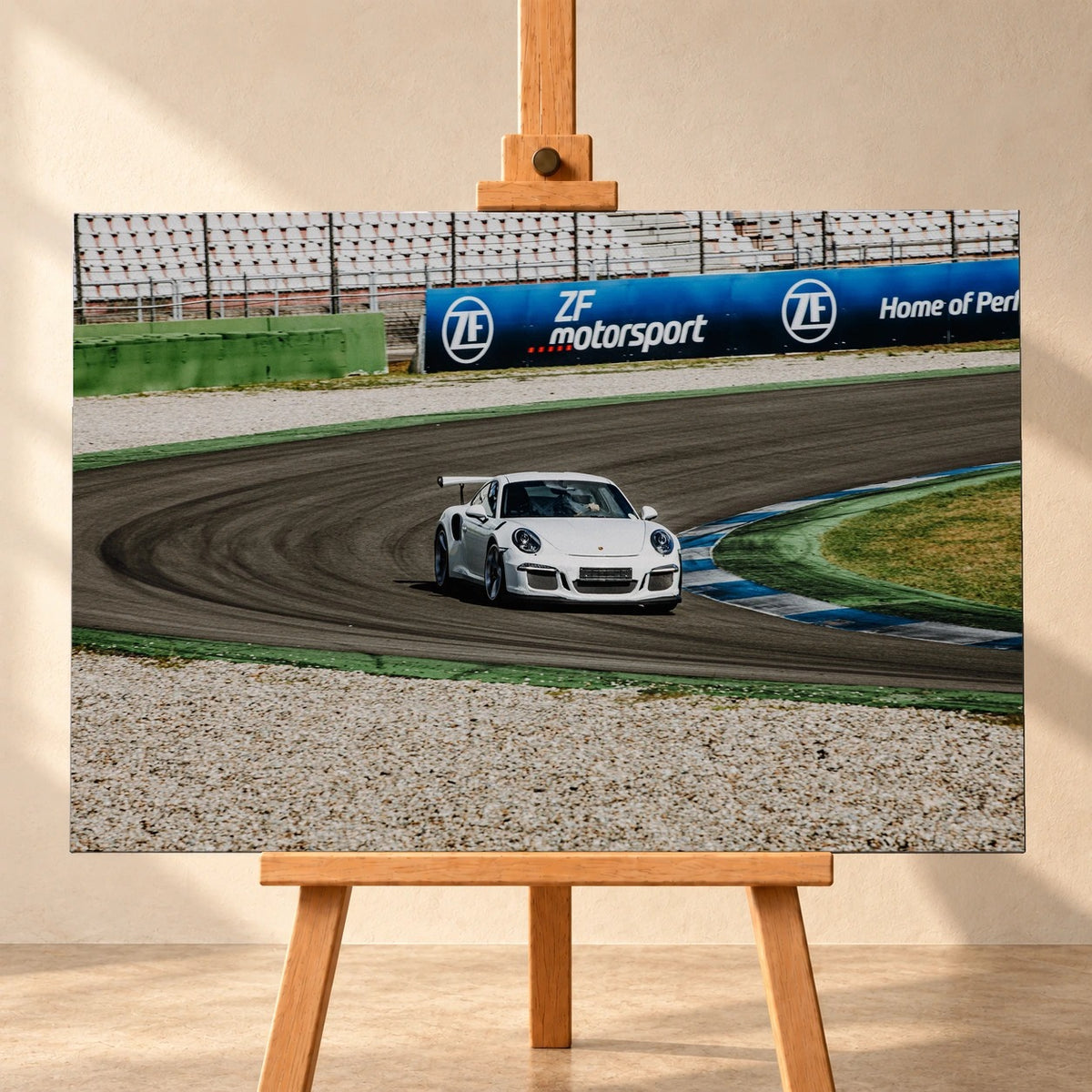 Frontale Visualisierung eines Leinwandbildes im Querformat auf einer Holzstaffelei vor heller Wand. Das Motiv zeigt einen weißen Porsche 911 mit großem Heckflügel in einer Rennstreckenkurve neben blau-weißen Randsteinen.