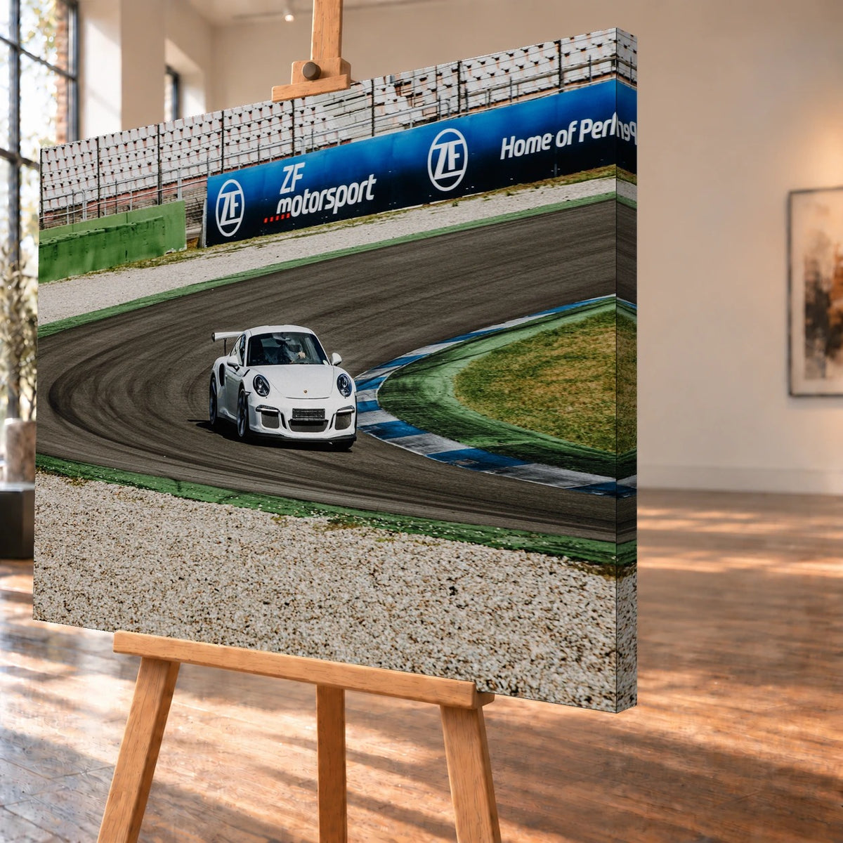 Leinwandbild als Visualisierung in schräger Ansicht auf einer Holzstaffelei, mit über die rechte Seitenkante fortgeführtem Motiv. Ein weißer Porsche 911 mit Heckflügel fährt durch eine Kurve; Reifenspuren und blau-weiße Randsteine prägen die Rennstrecke.