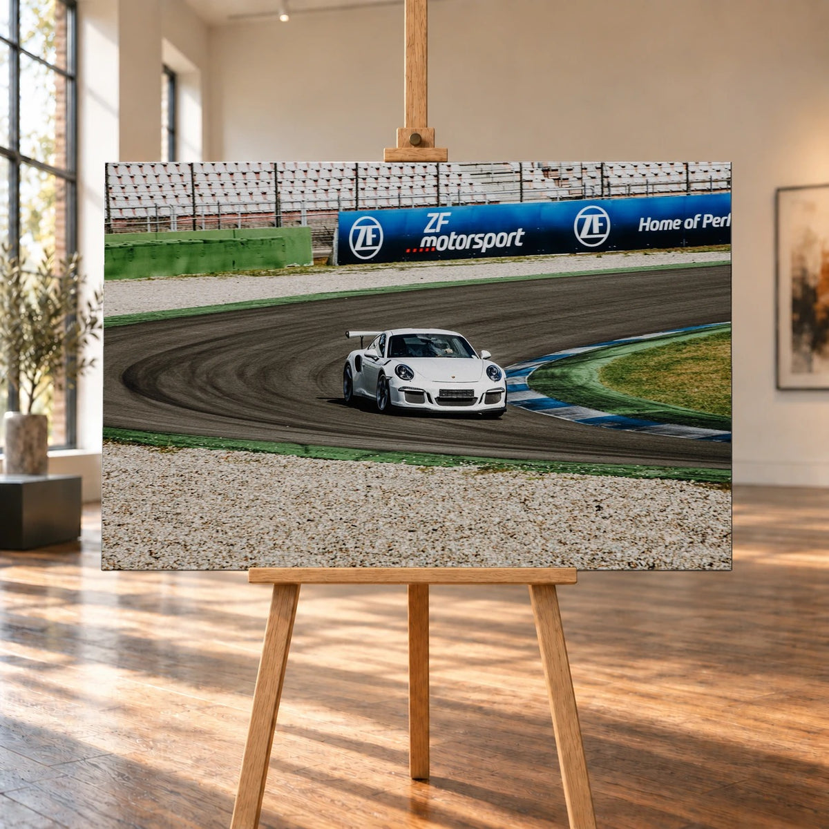 Raumvisualisierung eines querformatigen Leinwandbildes auf einer Holzstaffelei in einem hellen Raum mit hohen Fenstern und Holzboden. Auf dem Bild ist ein weißer Porsche 911 mit großem Heckflügel in einer Rennstreckenkurve zu sehen.