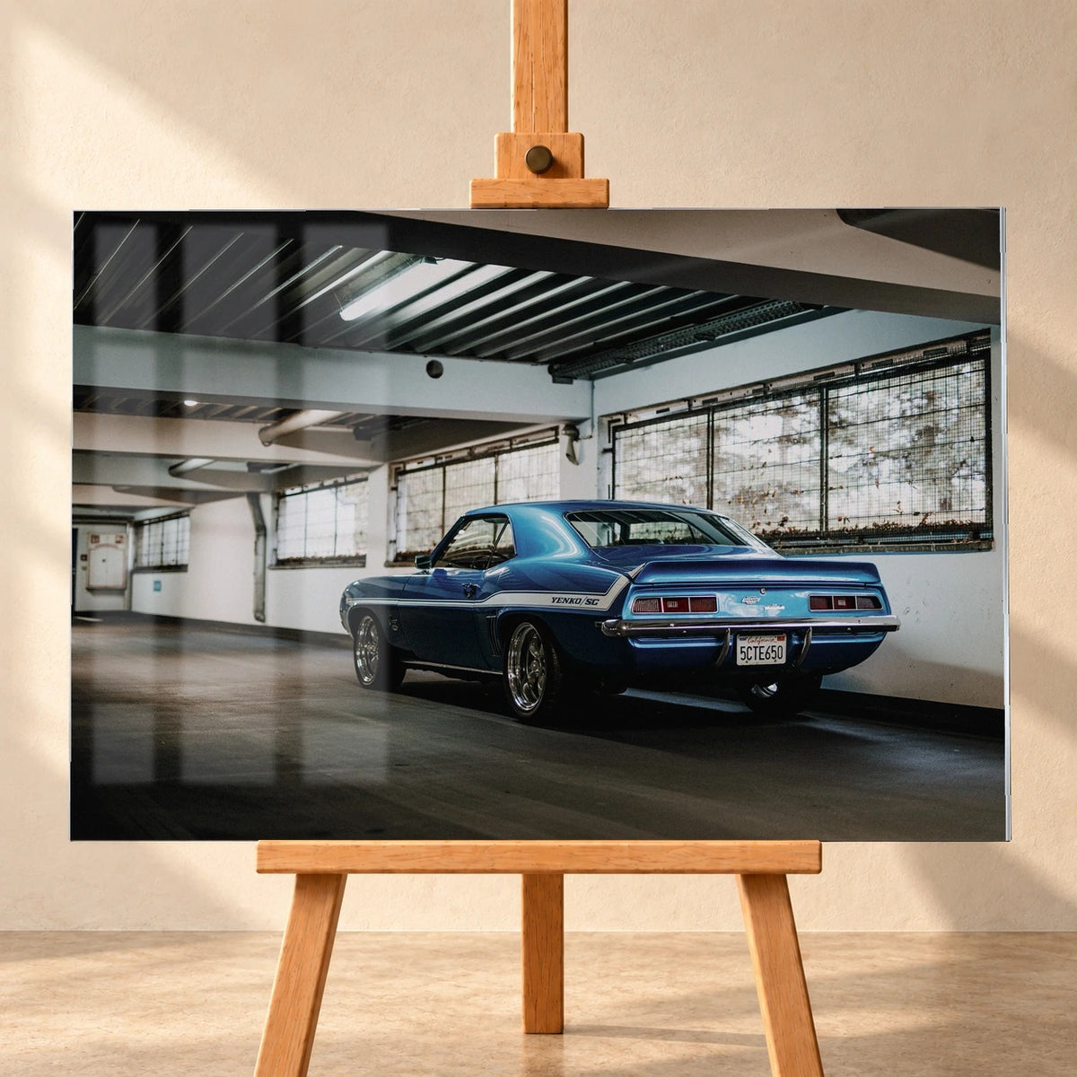 Frontal gezeigte Visualisierung eines Acrylglasbilds auf einer Holzstaffelei vor heller Wand. Sichtbare Fensterreflexionen überlagern das Motiv eines blauen Chevrolet Camaro Yenko in einer Halle, betrachtet von schräg hinten links.