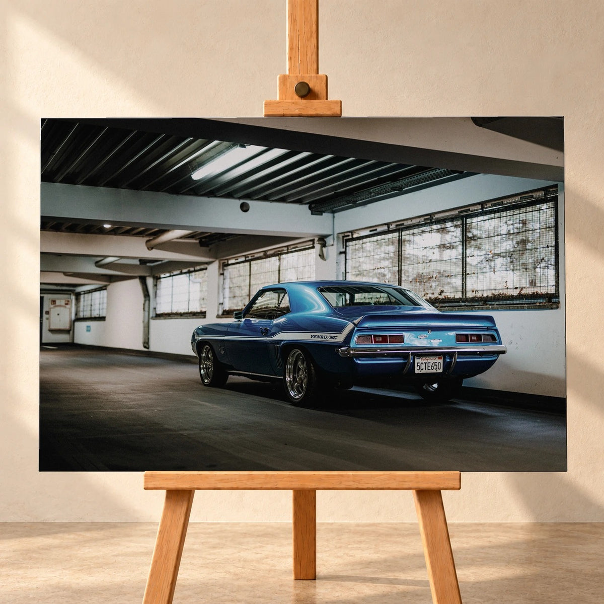 Visualisierung eines Leinwandbilds im Querformat auf einer Holzstaffelei vor heller Wand. Das Motiv zeigt einen blauen Chevrolet Camaro Yenko von schräg hinten links in einer Halle.