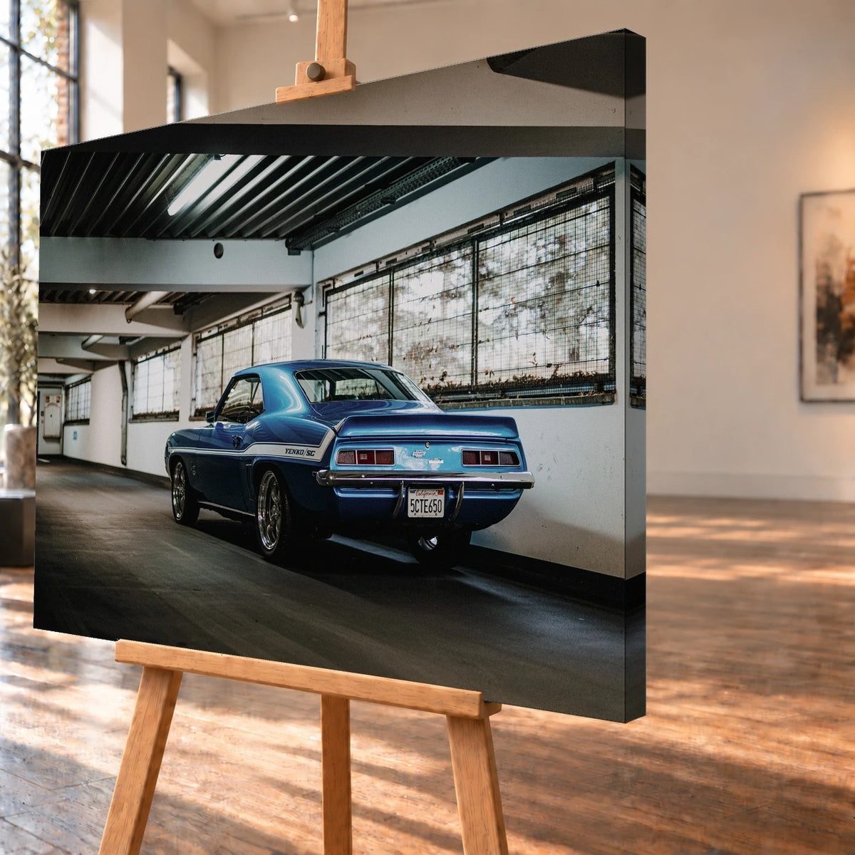 Schrägansicht eines visualisierten Leinwandbilds auf einer Holzstaffelei mit sichtbarer bedruckter Seitenkante. Im Motiv steht ein blauer Chevrolet Camaro Yenko mit weißem Seitenstreifen, von hinten links betrachtet, in einer Halle.