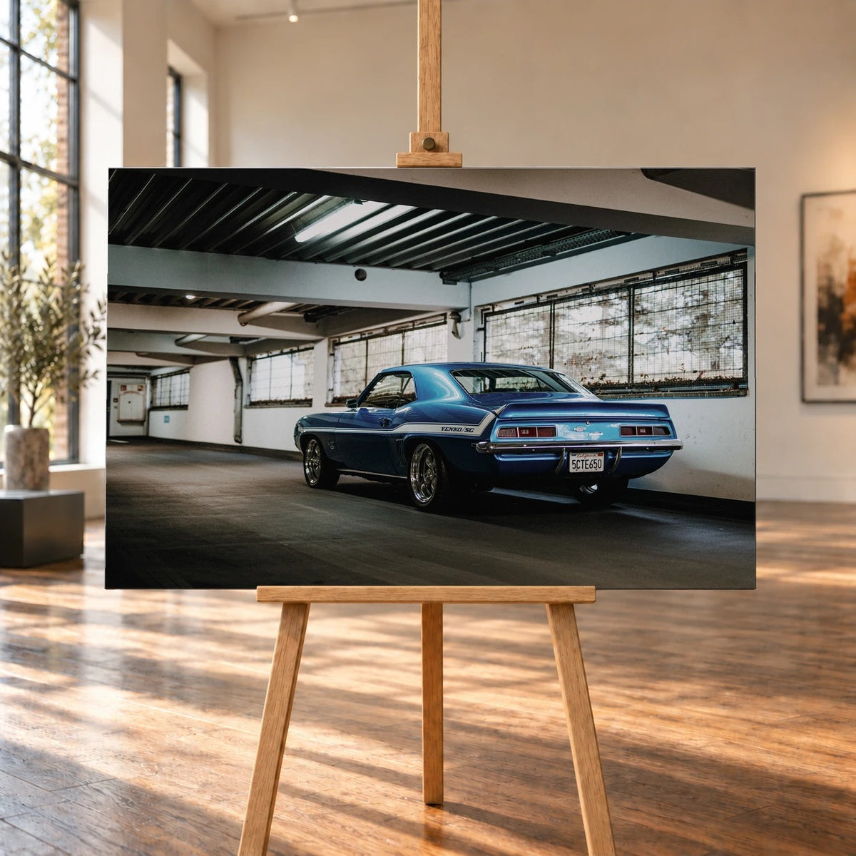 Raumvisualisierung mit einem querformatigen Leinwandbild auf einer Holzstaffelei in einem hellen Raum mit großen Fenstern und Holzboden. Das Bild zeigt einen blauen Chevrolet Camaro Yenko von hinten links in einer Halle.