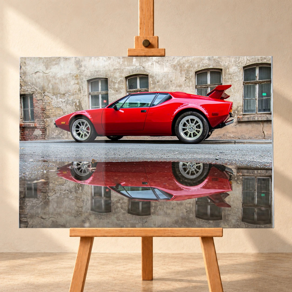 Ein roter De Tomaso Pantera GTS in Seitenansicht mit großem Heckflügel und Pfützenspiegelung vor einer verwitterten Fassade. Das Motiv ist als Acrylglas-Wandbild frontal auf einer Holzstaffelei vor heller Wand visualisiert.