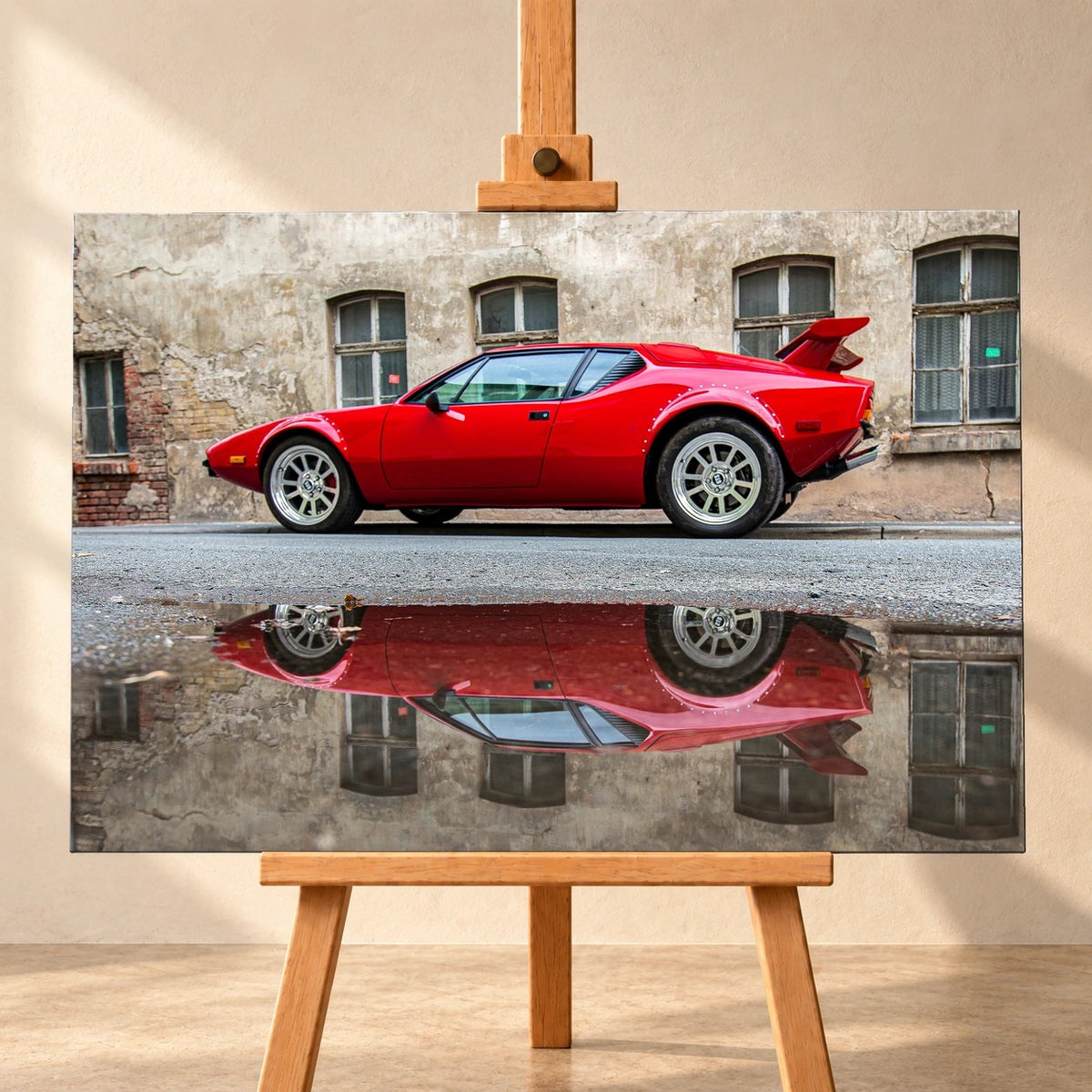 Visualisierung eines Leinwandbilds in frontaler Ansicht auf einer Holzstaffelei vor heller Wand. Das Motiv zeigt einen roten De Tomaso Pantera GTS seitlich vor einer verwitterten Fassade, mit seiner Spiegelung in einer Pfütze.