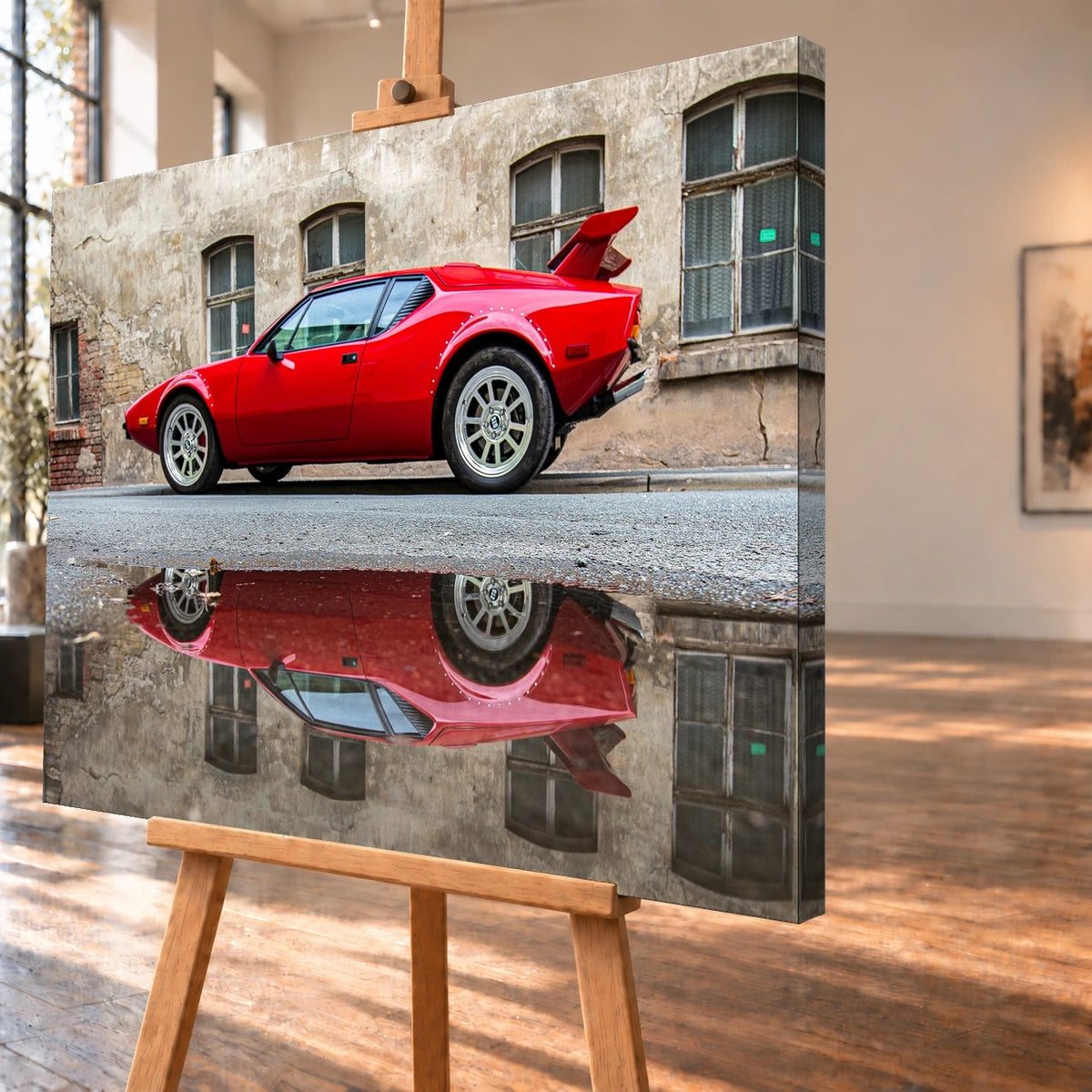 Schrägansicht eines visualisierten Leinwandbilds auf einer Holzstaffelei; das Motiv setzt sich über die sichtbare rechte Seitenkante fort. Zu sehen ist ein roter De Tomaso Pantera GTS vor einer verwitterten Fassade, gespiegelt im Wasser einer Pfütze.