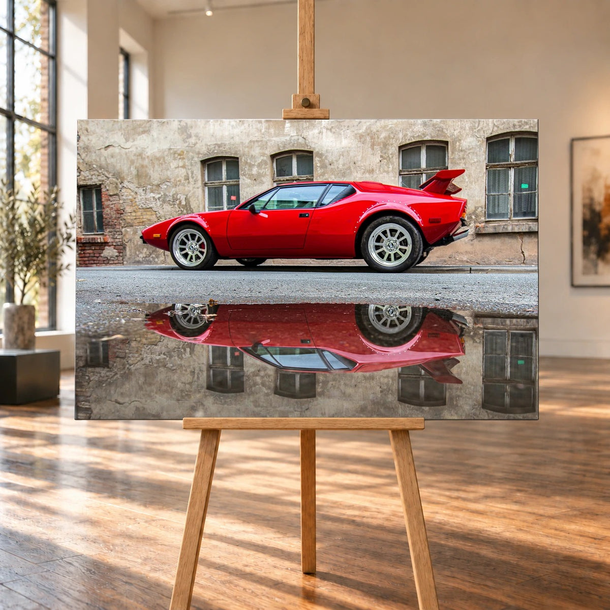Raumvisualisierung mit einem querformatigen Leinwandbild auf einer Holzstaffelei in einem hellen Raum mit großen Fenstern und Holzboden. Auf dem Bild steht ein roter De Tomaso Pantera GTS vor einer verwitterten Fassade und spiegelt sich in einer Pfütze.