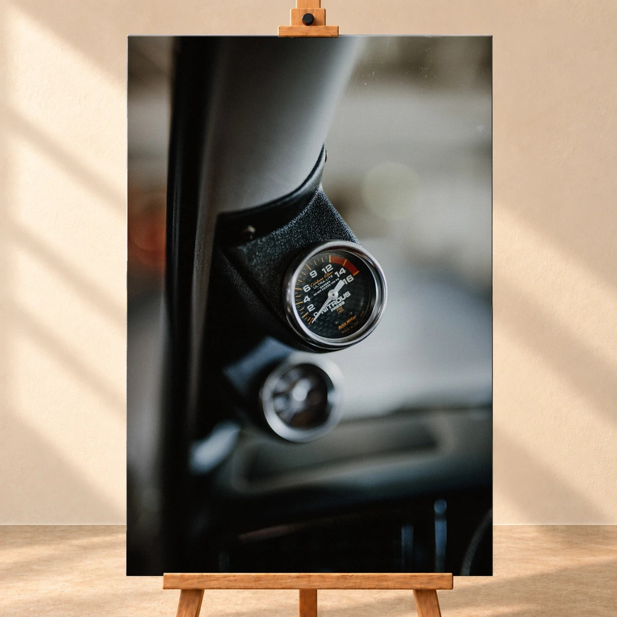 Visualisiertes Leinwandbild im Hochformat, frontal auf einer Holzstaffelei vor heller Wand. Das Motiv zeigt die silbern eingefasste Lachgas-Druckanzeige an der A-Säule von Jesse’s VW Jetta und eine unscharfe zweite Anzeige darunter.