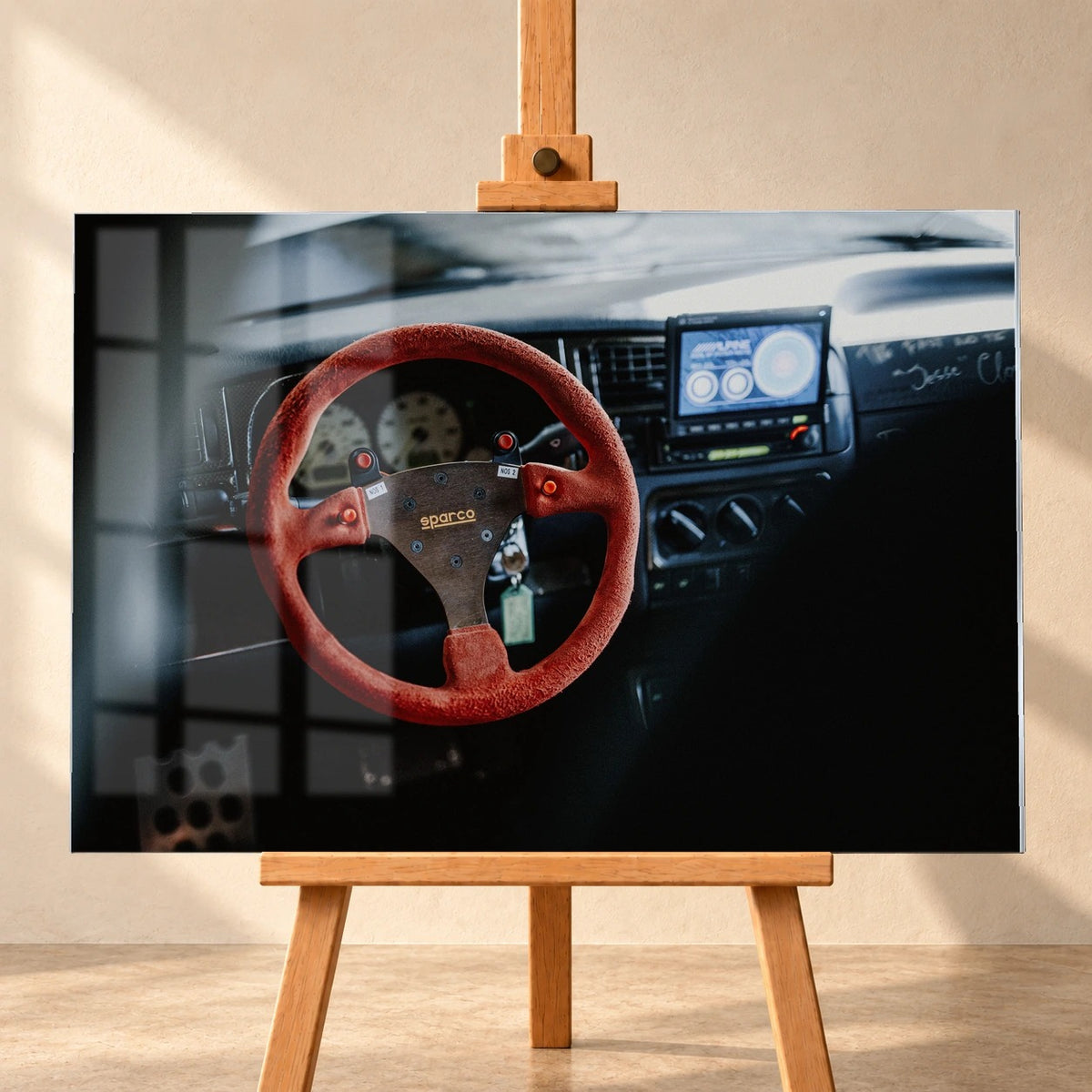Frontalansicht eines visualisierten Acrylglasbildes auf einer Holzstaffelei vor heller Wand, mit einer Fensterspiegelung auf der dargestellten Oberfläche. Das Motiv zeigt das rote Sparco-Lenkrad in Jesses VW Jetta aus „The Fast and the Furious“.