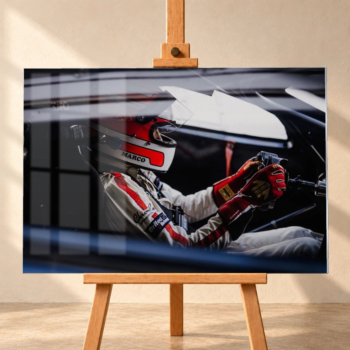 Visualisierung eines Acrylglasbilds im Querformat auf einer Holzstaffelei vor heller Wand. Eine Fensterspiegelung überlagert links das Motiv eines Rennfahrers mit rot-weißem Helm, hellem Rennanzug und roten Handschuhen am Lenkrad.