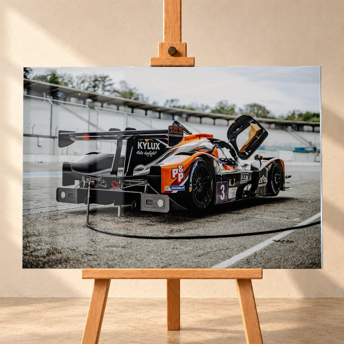 Frontalansicht eines querformatigen Acrylglas-Wandbilds als Visualisierung auf einer Holzstaffelei vor heller Wand. Das Motiv zeigt einen orange-schwarz-weißen Rennwagen mit Startnummer 3, breitem Heckflügel und hochgeklappter Tür in der Boxengasse.