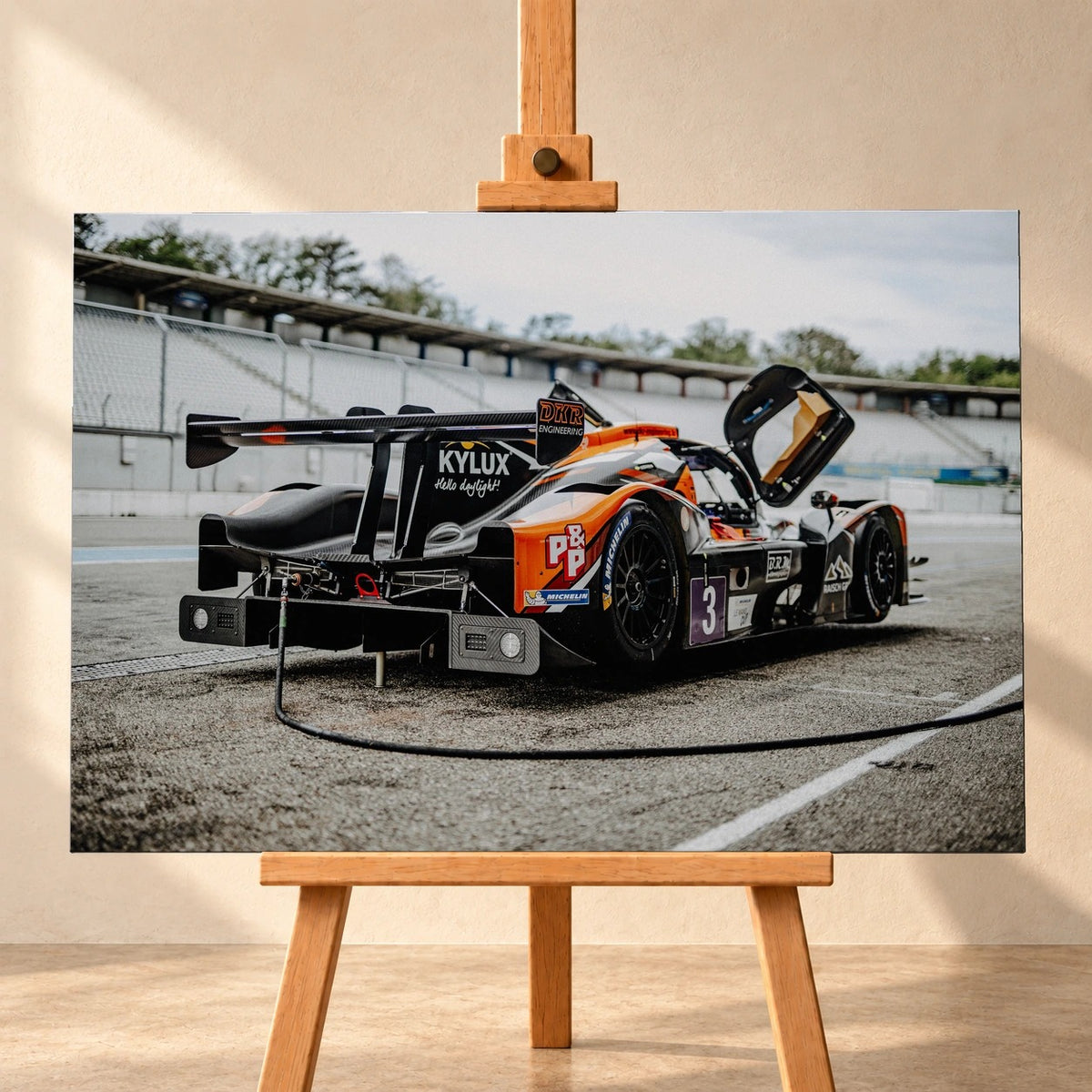 Visualisierung einer querformatigen Leinwand auf einer Holzstaffelei vor heller Wand. Das Motiv zeigt einen orange-schwarz-weißen Rennwagen mit Startnummer 3 schräg von hinten, mit großem Heckflügel und hochgeklappter Tür.