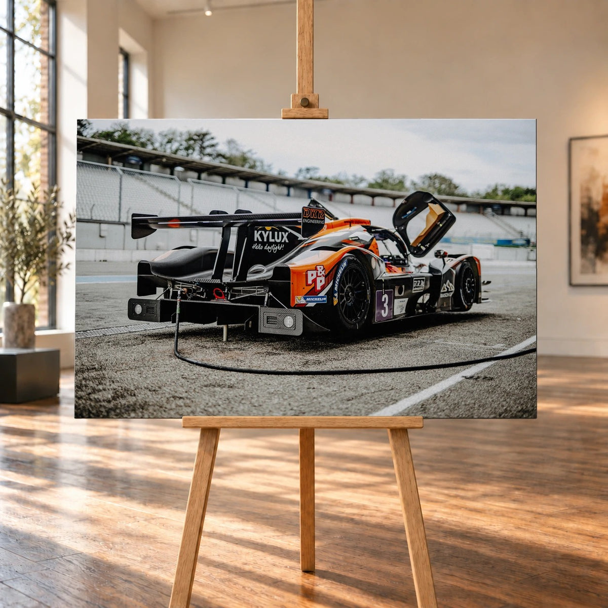 Raumvisualisierung einer querformatigen Leinwand auf einer Holzstaffelei in einem hellen Raum mit hohen Fenstern und Holzboden. Darauf ein orange-schwarz-weißer Rennwagen mit Startnummer 3 und hochgeklappter Tür, schräg von hinten gesehen.