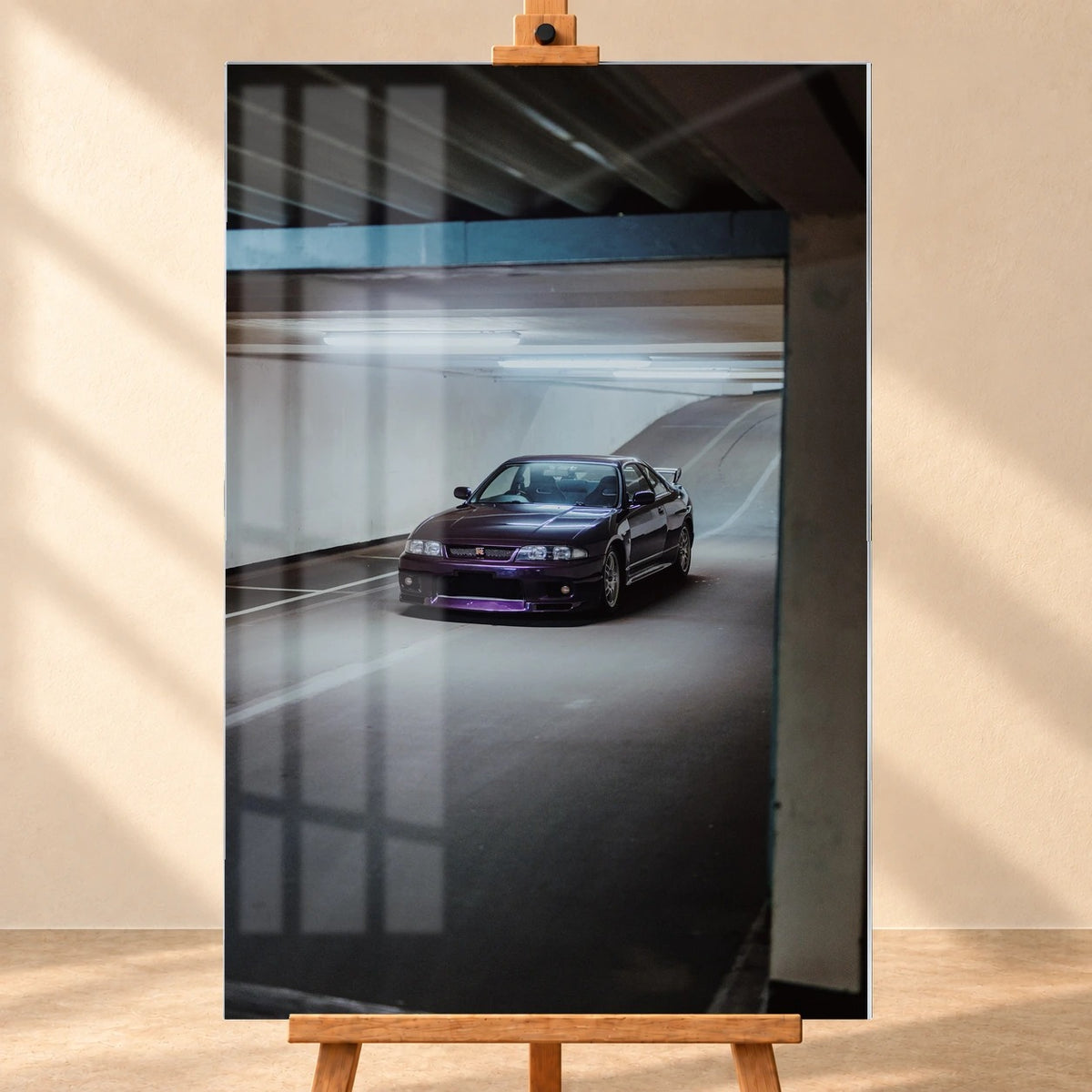 Visualisierung eines Acrylglasbildes im Hochformat, frontal auf einer Holzstaffelei vor beiger Wand. Fensterspiegelungen überlagern das Motiv eines dunkelvioletten Nissan Skyline GT-R R33 auf einer beleuchteten Parkhausrampе.