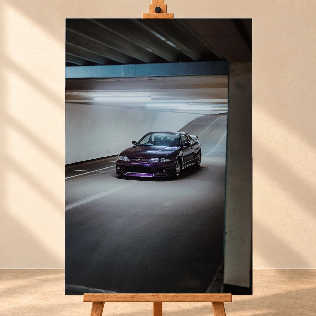 Frontale Visualisierung eines Leinwandbildes im Hochformat auf einer Holzstaffelei vor beiger Wand. Das Motiv zeigt einen dunkelvioletten Nissan Skyline GT-R R33 schräg von vorn auf einer beleuchteten Parkhausrampе.