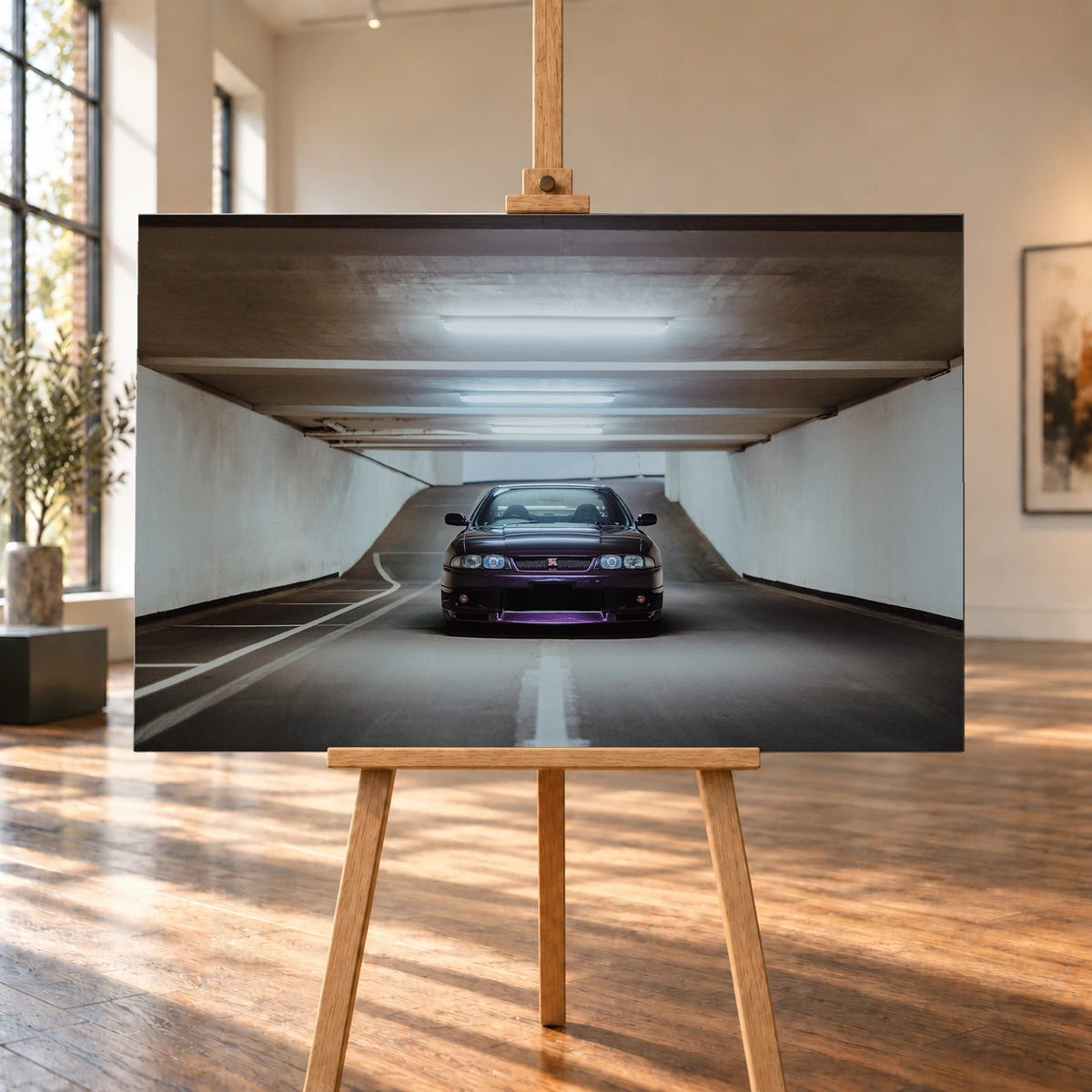 Raumvisualisierung mit einem querformatigen Leinwandbild auf einer Holzstaffelei in einem hellen Raum mit großen Fenstern und Holzboden. Das Bild zeigt einen dunkelvioletten Nissan Skyline GT-R R33 von vorn in einer beleuchteten Tiefgaragenzufahrt.