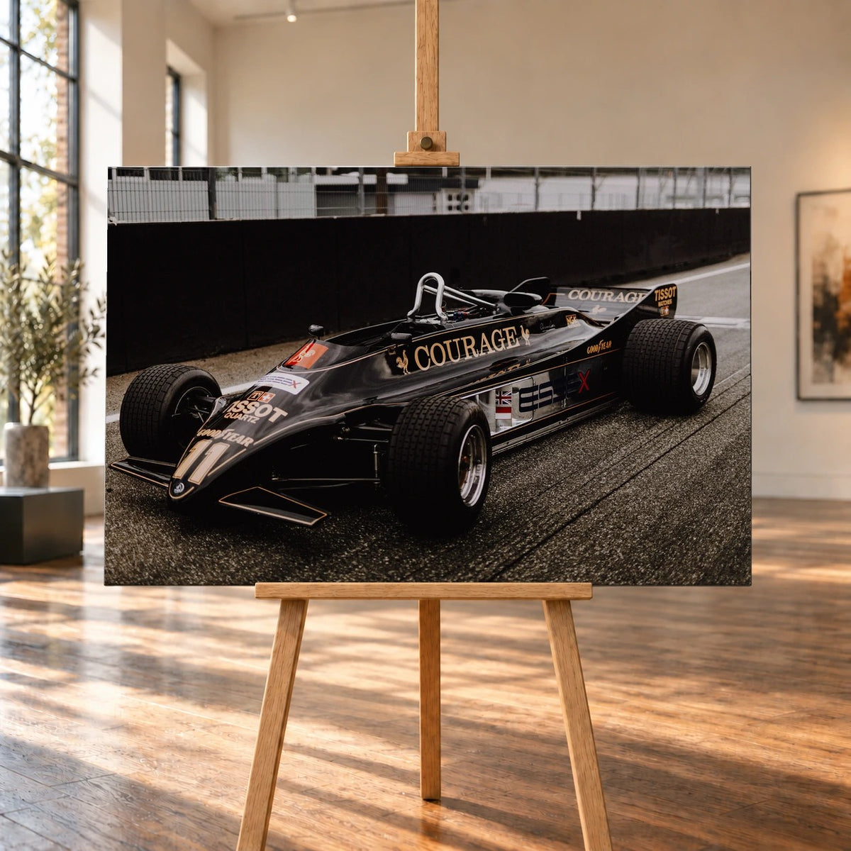 Raumvisualisierung eines querformatigen Leinwandbilds auf einer Holzstaffelei in einem hellen Raum mit hohen Fenstern und Holzboden. Das Bild zeigt den schwarzen Lotus 88 schräg von vorn, mit goldenen Linien, COURAGE-Schriftzug und freistehenden Rädern.