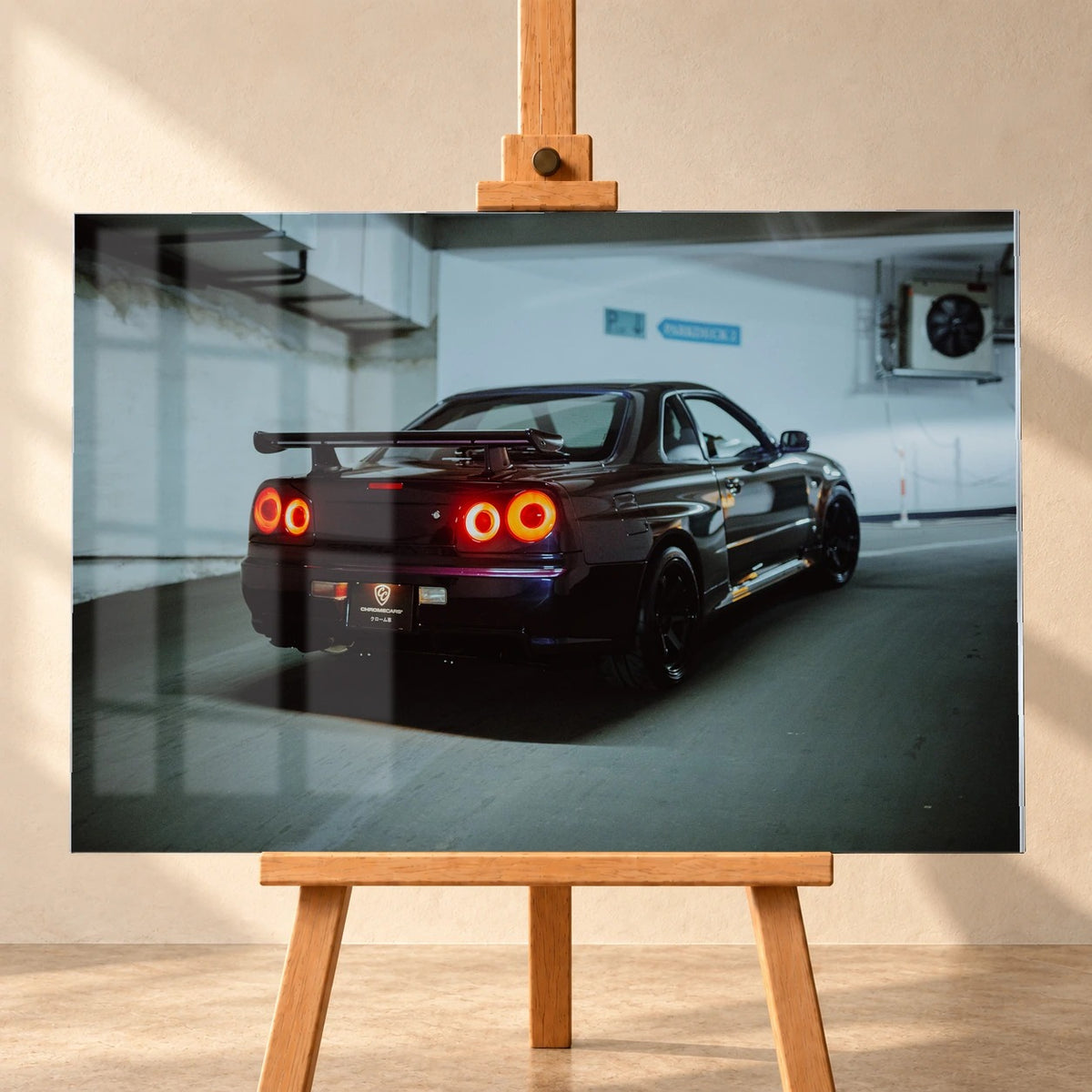 Visualisierung eines Acrylglasbilds im Querformat, frontal auf einer Holzstaffelei vor heller Wand. Fensterreflexionen überlagern links das Motiv eines dunkelvioletten Nissan Skyline R34, der schräg von hinten mit großem Heckflügel und vier roten runden Rücklichtern in einer Tiefgarage zu sehen ist.