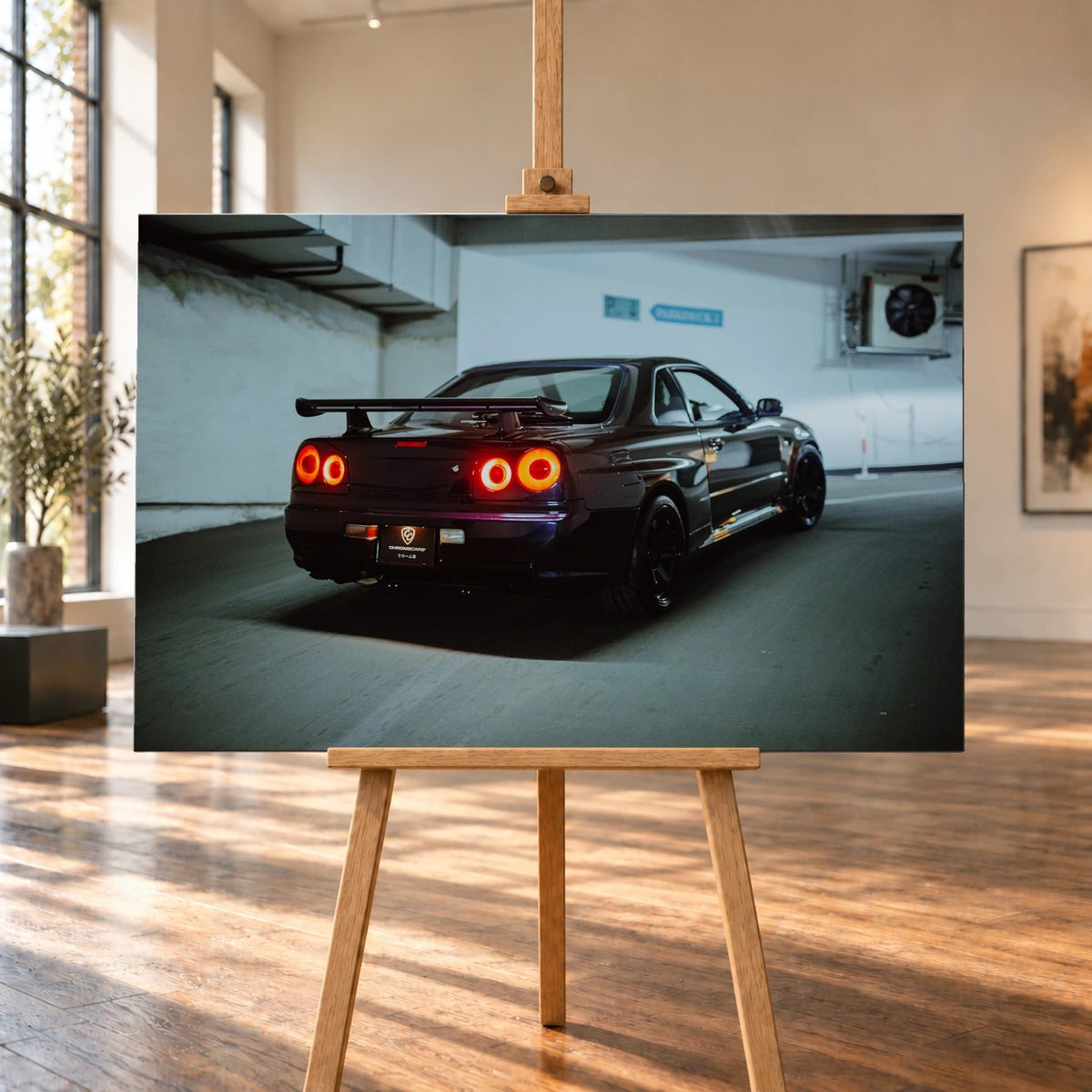 Raumvisualisierung mit einem querformatigen Leinwandbild auf einer Holzstaffelei in einem hellen Raum mit hohen Fenstern und Holzboden. Zu sehen ist ein dunkelvioletter Nissan Skyline R34 in Heck-Dreiviertelansicht, mit leuchtenden runden Rücklichtern in einer Tiefgarage.