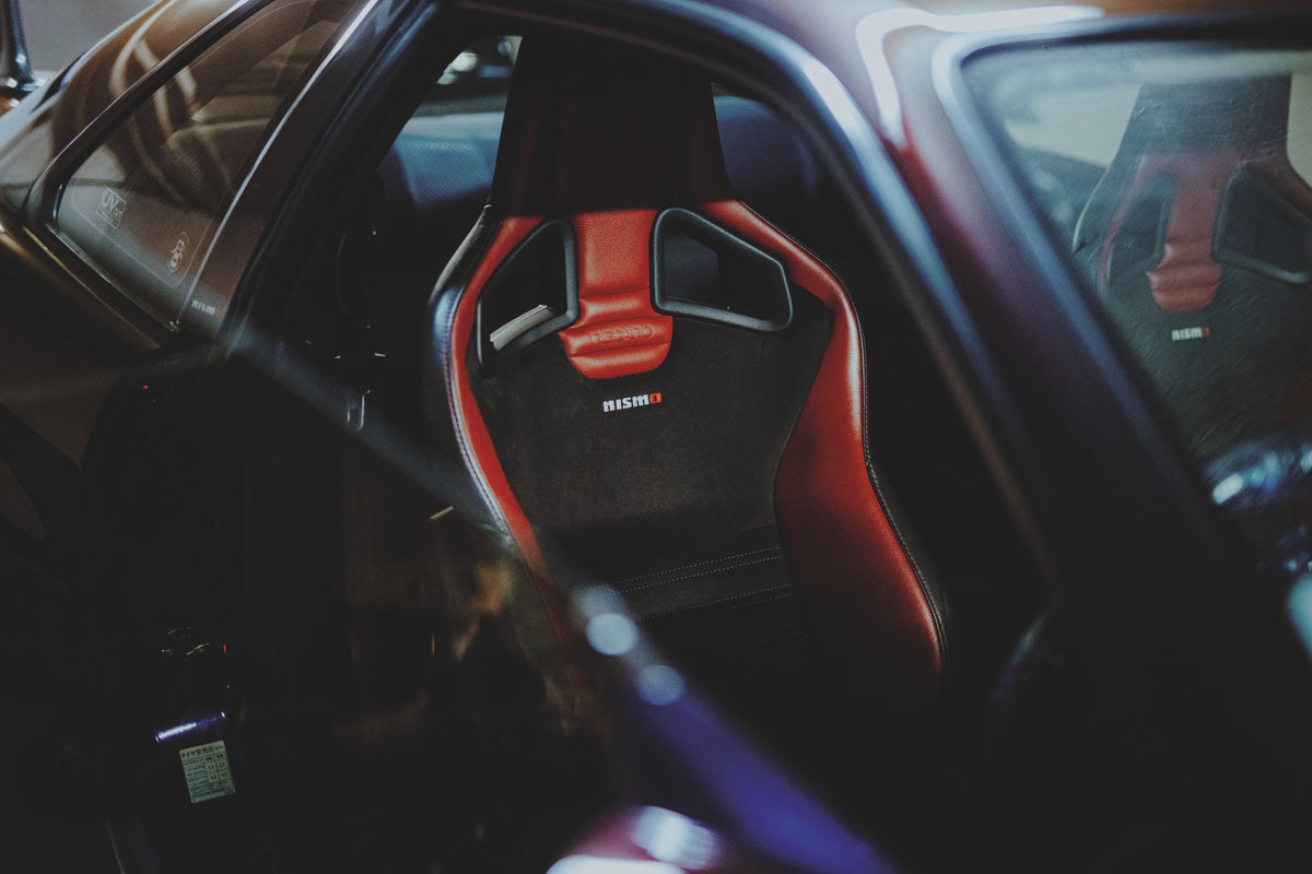 Geöffnete Tür eines Nissan Skyline R34 mit Blick auf einen schwarz-roten Sportsitz und den dunklen Innenraum.