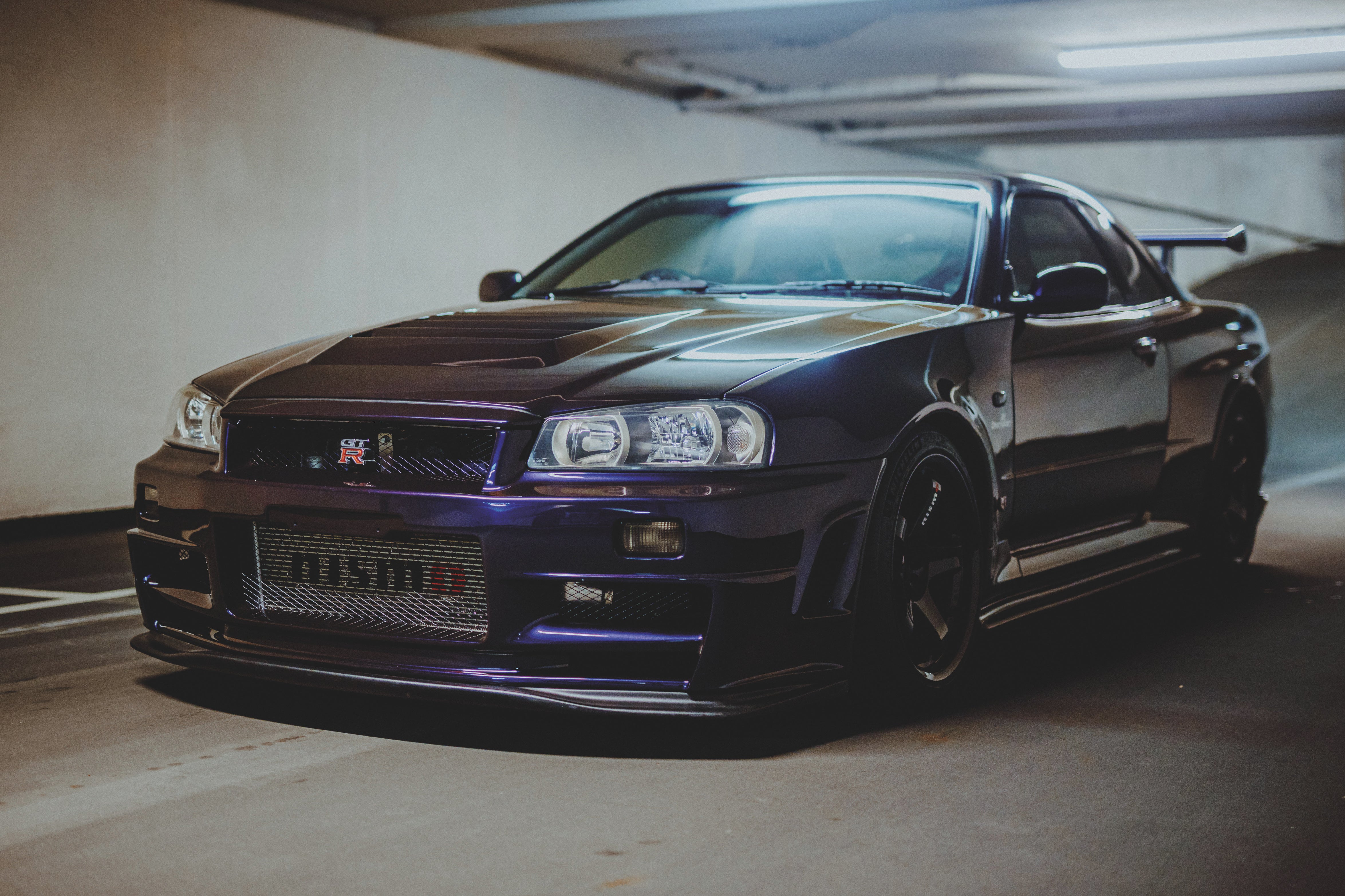 Dunkler Nissan Skyline R34 schräg von vorn unter Deckenleuchten in einer Tiefgarage.