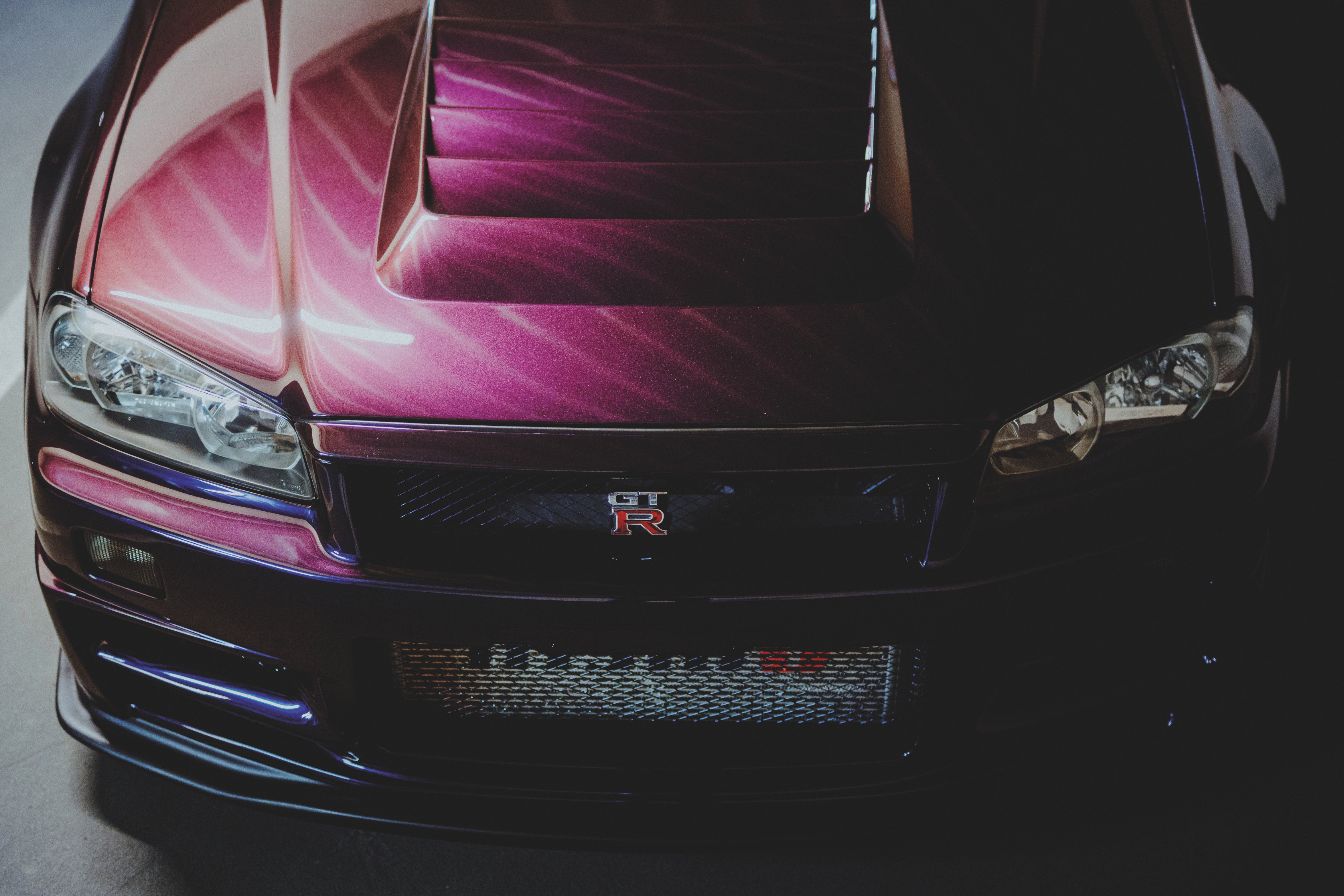 Breite Nahansicht der Front eines Nissan Skyline R34 mit violett reflektierender Motorhaube, Scheinwerfern und GT-R-Emblem.