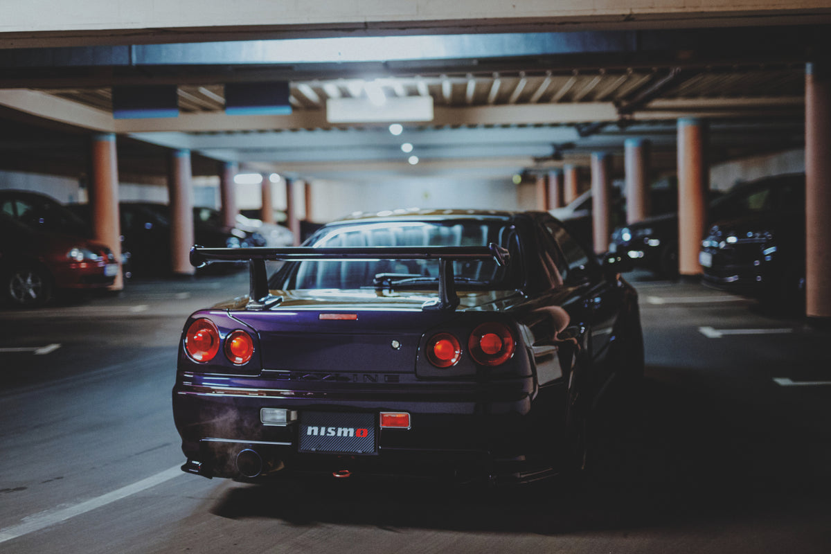 Nissan Skyline R34 schräg von hinten in einer Garage mit mehreren sichtbaren Stellplätzen und Deckenbalken.