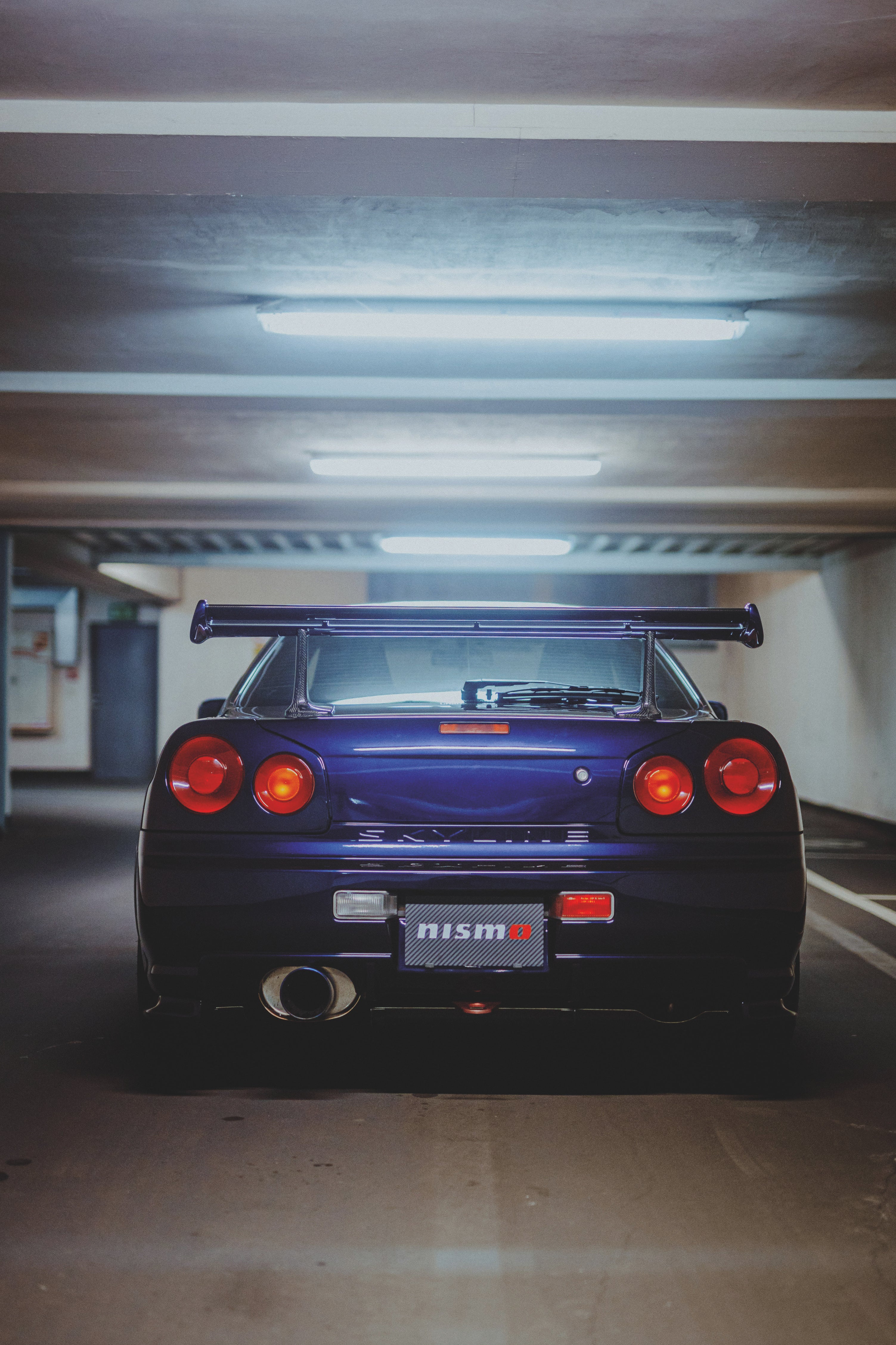 Gerade Heckansicht eines violetten Nissan Skyline R34 mit vier runden Rückleuchten unter Deckenleuchten.