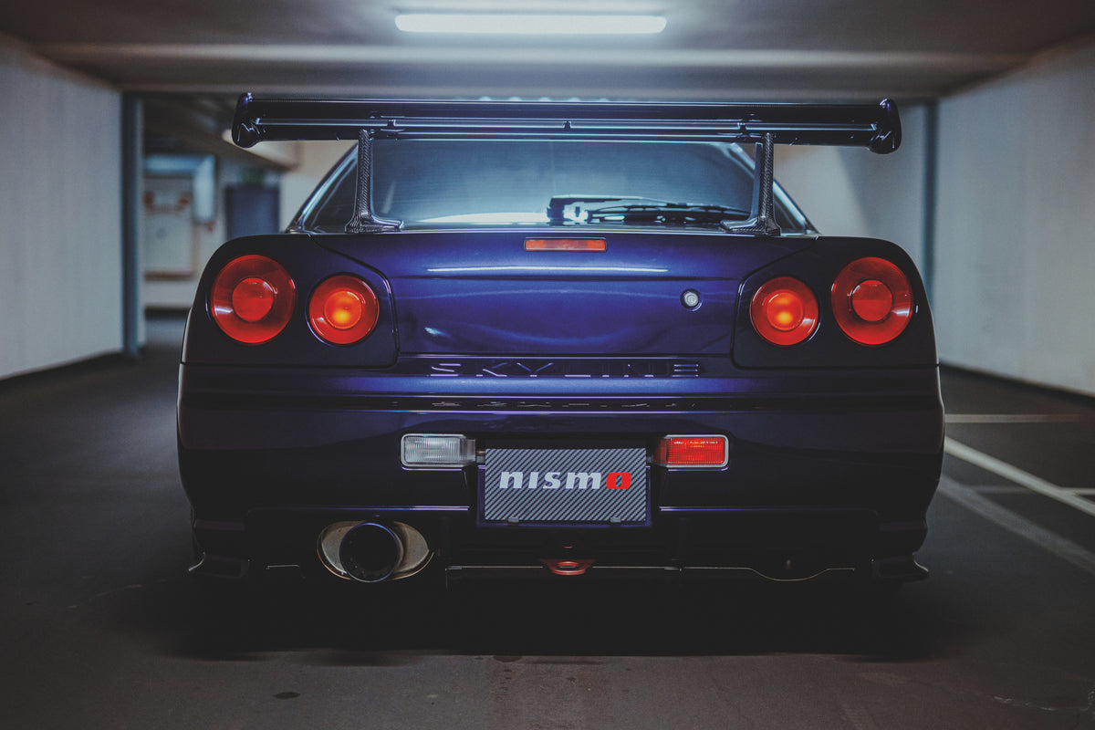 Gerade Heckansicht eines violetten Nissan Skyline R34 mit vier runden Rückleuchten, Heckflügel und großem Auspuffendrohr.