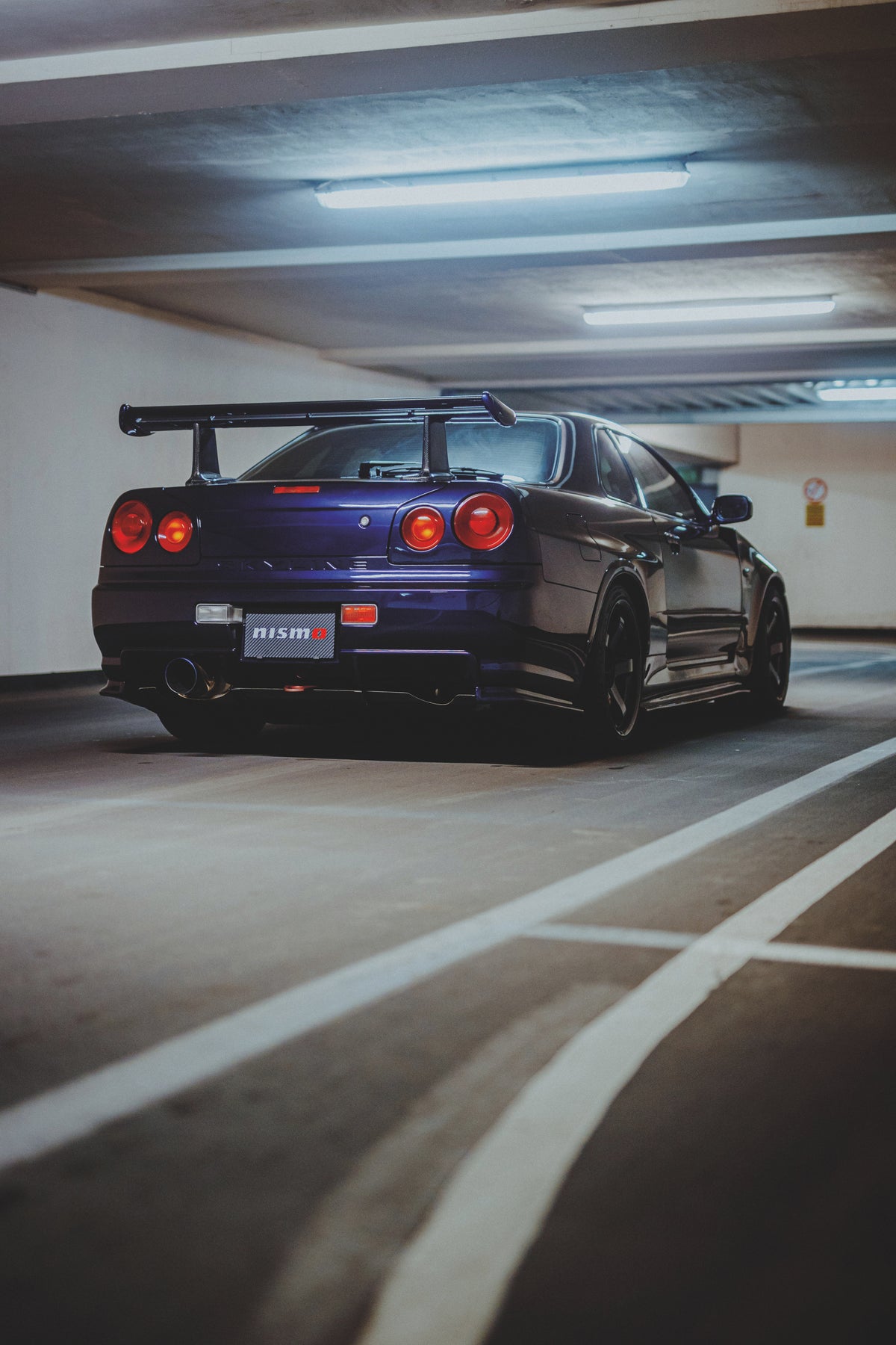 Nissan Skyline R34 schräg von hinten neben weißen Bodenmarkierungen, mit Heckflügel und runden Rückleuchten.