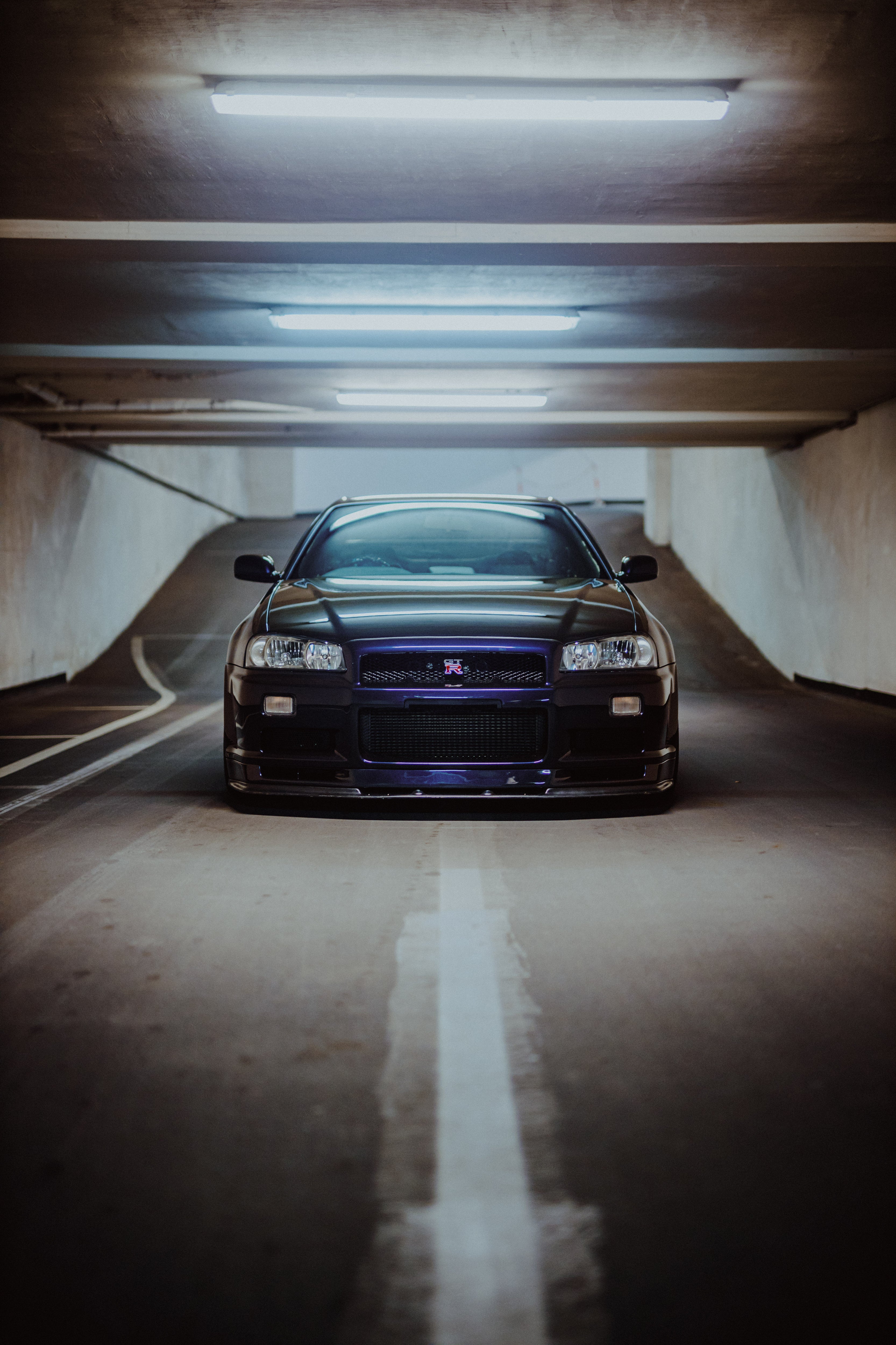 Nissan Skyline R34 frontal und mittig über einer weißen Bodenlinie unter Garagenleuchten.
