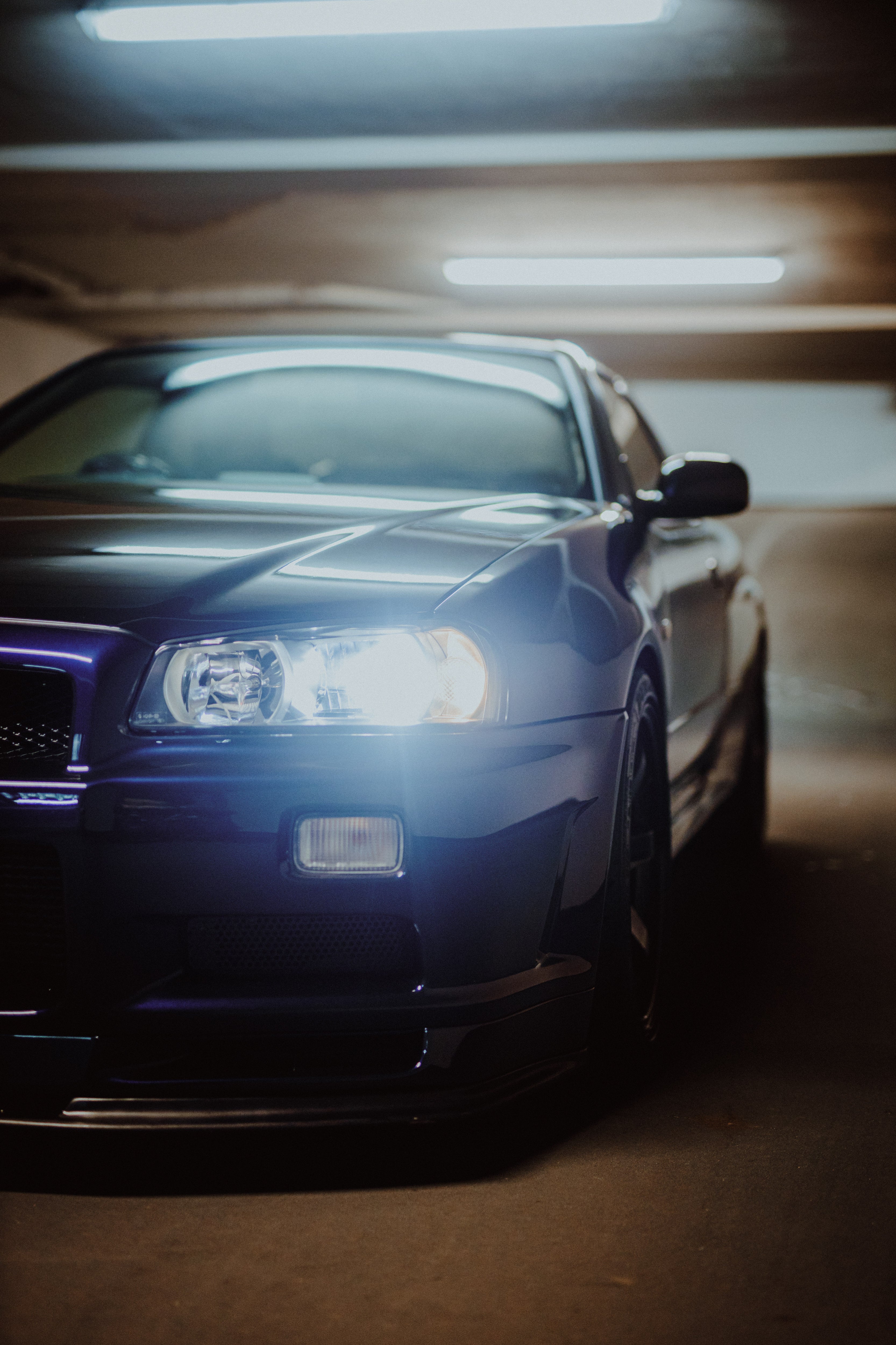Nahe schräge Frontansicht eines Nissan Skyline R34 mit hell leuchtendem Scheinwerfer in einer Tiefgarage.