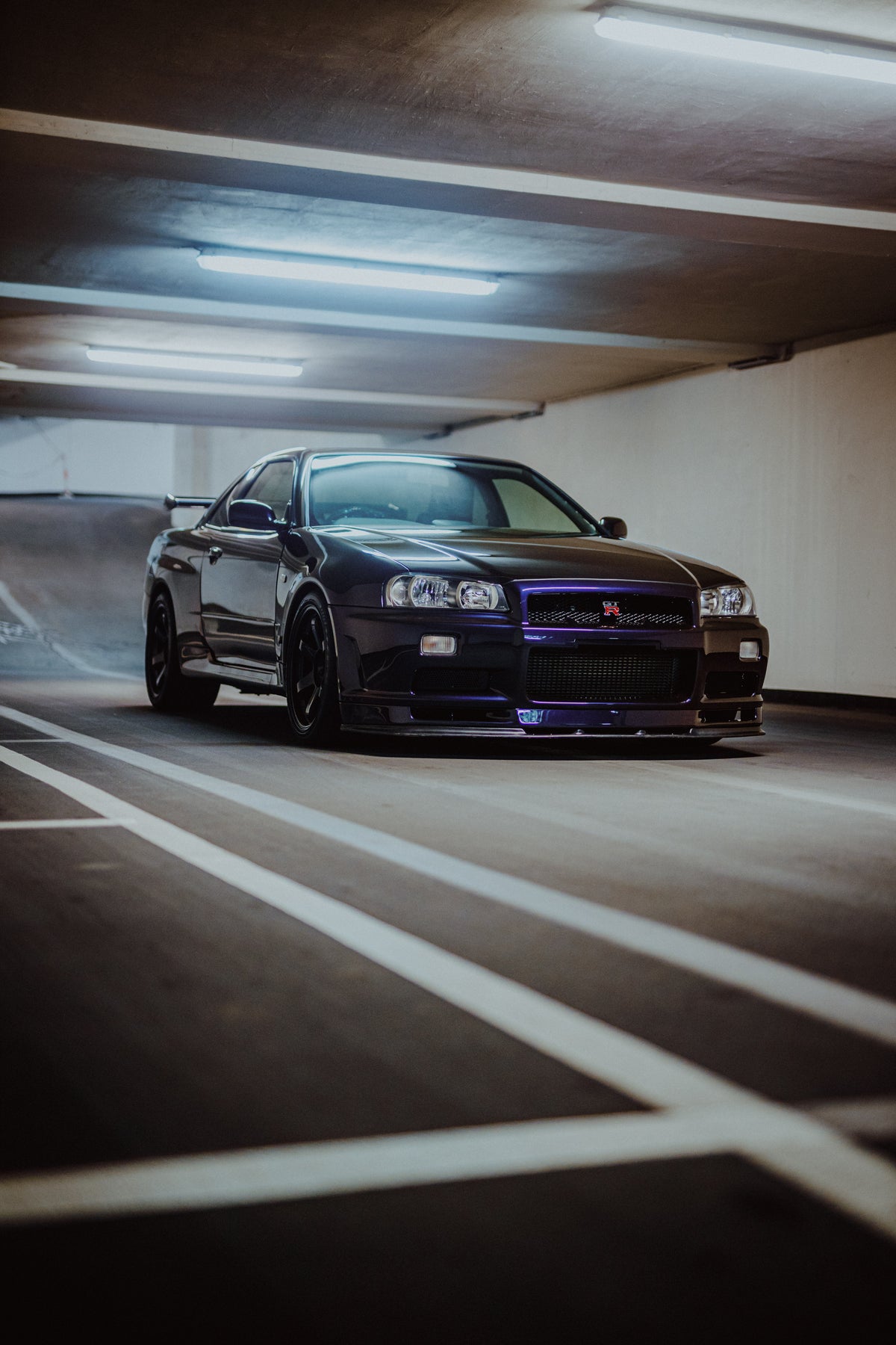 Dunkelvioletter Nissan Skyline R34 schräg von vorn hinter weißen Bodenlinien in einer Tiefgarage.