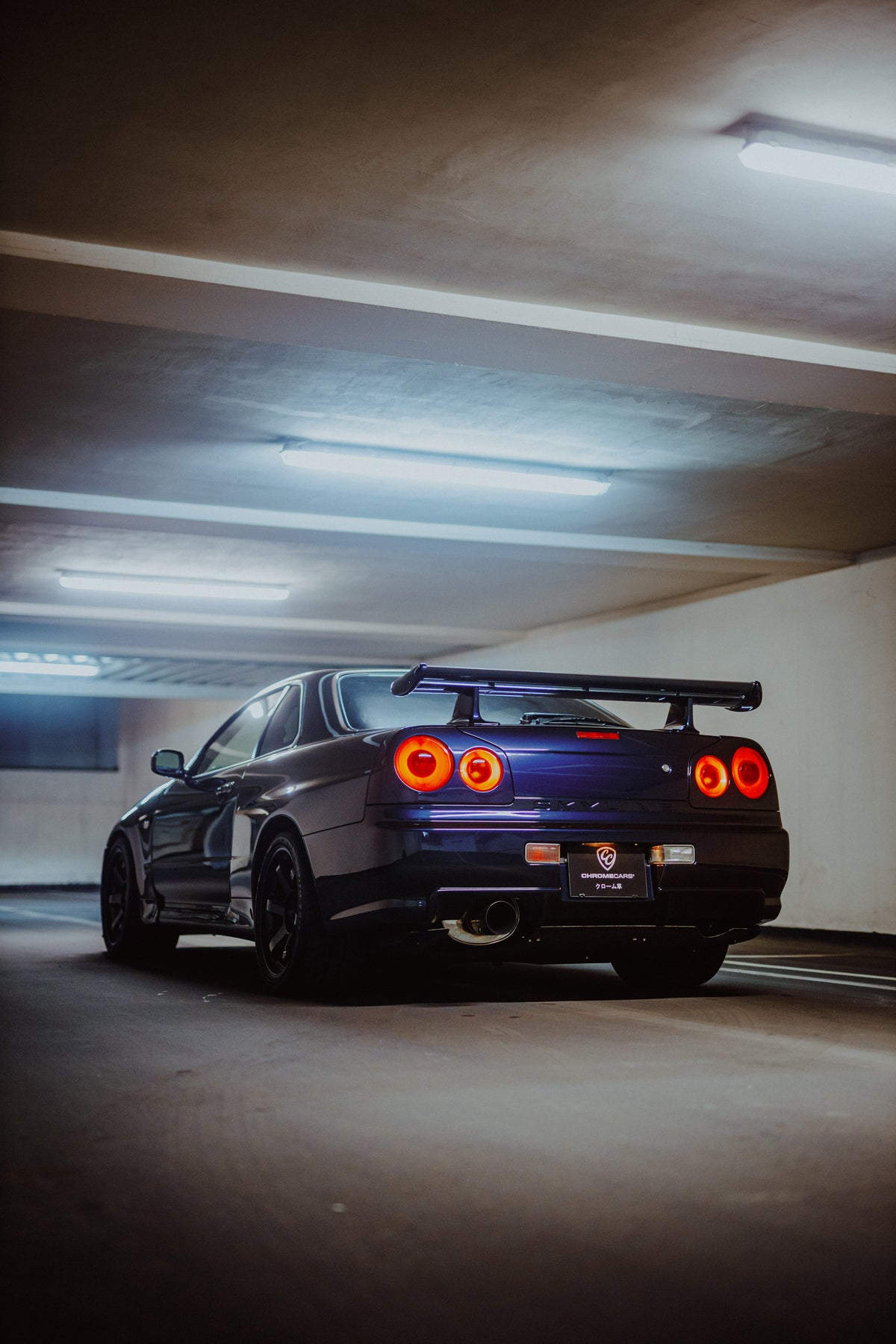 Dunkler Nissan Skyline R34 schräg von hinten unter Garagenleuchten mit vier runden roten Rückleuchten.
