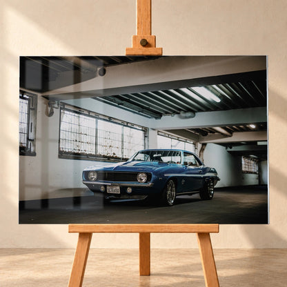 Der Camaro Yenko steht schräg von vorn in einer alten, lichtdurchfluteten Halle; Fensterreihen und Beton schaffen industrielle Atmosphäre. Motiv „Silver Pulse 2“ als Acrylglas-Wandbild, frontal auf einer Holzstaffelei.