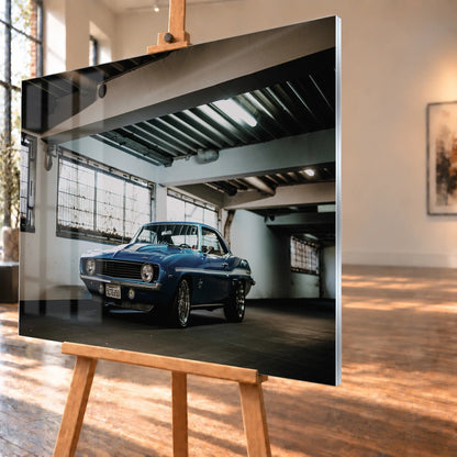 Der Camaro Yenko steht schräg von vorn in einer alten, lichtdurchfluteten Halle; Fensterreihen und Beton schaffen industrielle Atmosphäre. Motiv „Silver Pulse 2“ als Acrylglas-Wandbild, schräg von der Seite auf einer Holzstaffelei.