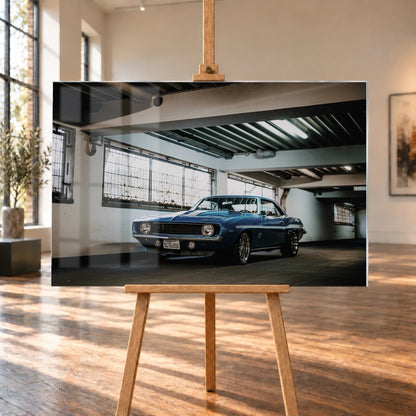 Der Camaro Yenko steht schräg von vorn in einer alten, lichtdurchfluteten Halle; Fensterreihen und Beton schaffen industrielle Atmosphäre. Motiv „Silver Pulse 2“ als Acrylglas-Wandbild, auf einer Holzstaffelei in einem hellen Galerieraum.