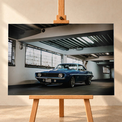 Der Camaro Yenko steht schräg von vorn in einer alten, lichtdurchfluteten Halle; Fensterreihen und Beton schaffen industrielle Atmosphäre. Motiv „Silver Pulse 2“ als Alu gebürstet-Wandbild, frontal auf einer Holzstaffelei.