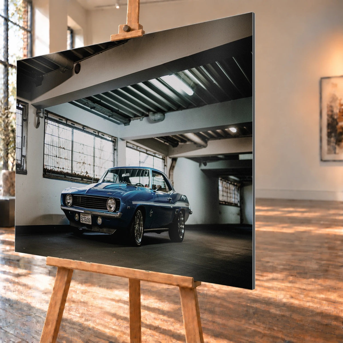 Der Camaro Yenko steht schräg von vorn in einer alten, lichtdurchfluteten Halle; Fensterreihen und Beton schaffen industrielle Atmosphäre. Motiv „Silver Pulse 2“ als Alu gebürstet-Wandbild, schräg von der Seite auf einer Holzstaffelei.
