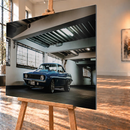 Der Camaro Yenko steht schräg von vorn in einer alten, lichtdurchfluteten Halle; Fensterreihen und Beton schaffen industrielle Atmosphäre. Motiv „Silver Pulse 2“ als Alu gebürstet-Wandbild, schräg von der Seite auf einer Holzstaffelei.