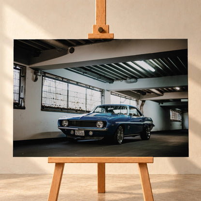 Der Camaro Yenko steht schräg von vorn in einer alten, lichtdurchfluteten Halle; Fensterreihen und Beton schaffen industrielle Atmosphäre. Motiv „Silver Pulse 2“ als Leinwand-Wandbild, frontal auf einer Holzstaffelei.