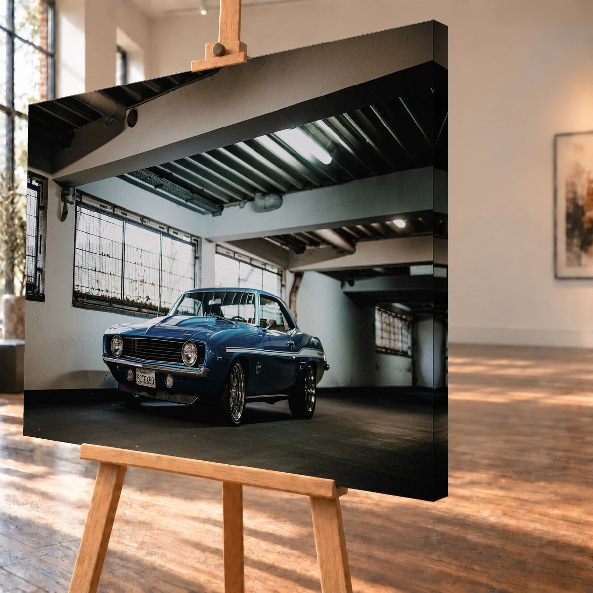 Der Camaro Yenko steht schräg von vorn in einer alten, lichtdurchfluteten Halle; Fensterreihen und Beton schaffen industrielle Atmosphäre. Motiv „Silver Pulse 2“ als Leinwand-Wandbild, schräg von der Seite auf einer Holzstaffelei.