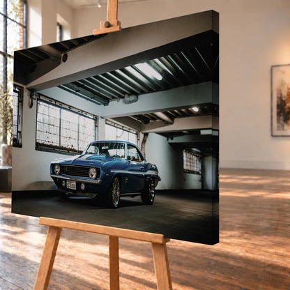 Der Camaro Yenko steht schräg von vorn in einer alten, lichtdurchfluteten Halle; Fensterreihen und Beton schaffen industrielle Atmosphäre. Motiv „Silver Pulse 2“ als Leinwand-Wandbild, schräg von der Seite auf einer Holzstaffelei.