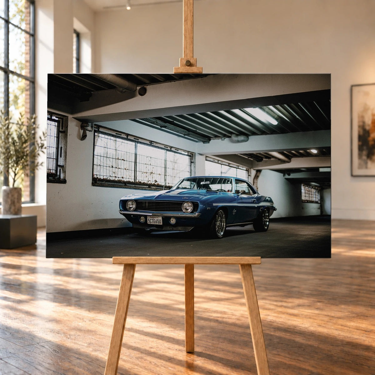 Der Camaro Yenko steht schräg von vorn in einer alten, lichtdurchfluteten Halle; Fensterreihen und Beton schaffen industrielle Atmosphäre. Motiv „Silver Pulse 2“ als Leinwand-Wandbild, auf einer Holzstaffelei in einem hellen Galerieraum.