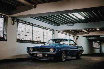 Der Camaro Yenko steht schräg von vorn in einer alten, lichtdurchfluteten Halle; Fensterreihen und Beton schaffen industrielle Atmosphäre. Originalmotiv „Silver Pulse 2“ im Querformat.