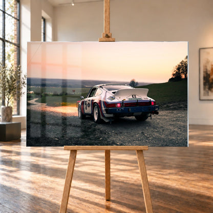 Der Porsche 911 Safari steht von hinten auf einer Anhöhe im Abendrot; weiter Himmel und orangefarbene Horizontlinie öffnen das Bild. Motiv „Fast Motion“ als Acrylglas-Wandbild, auf einer Holzstaffelei in einem hellen Galerieraum.