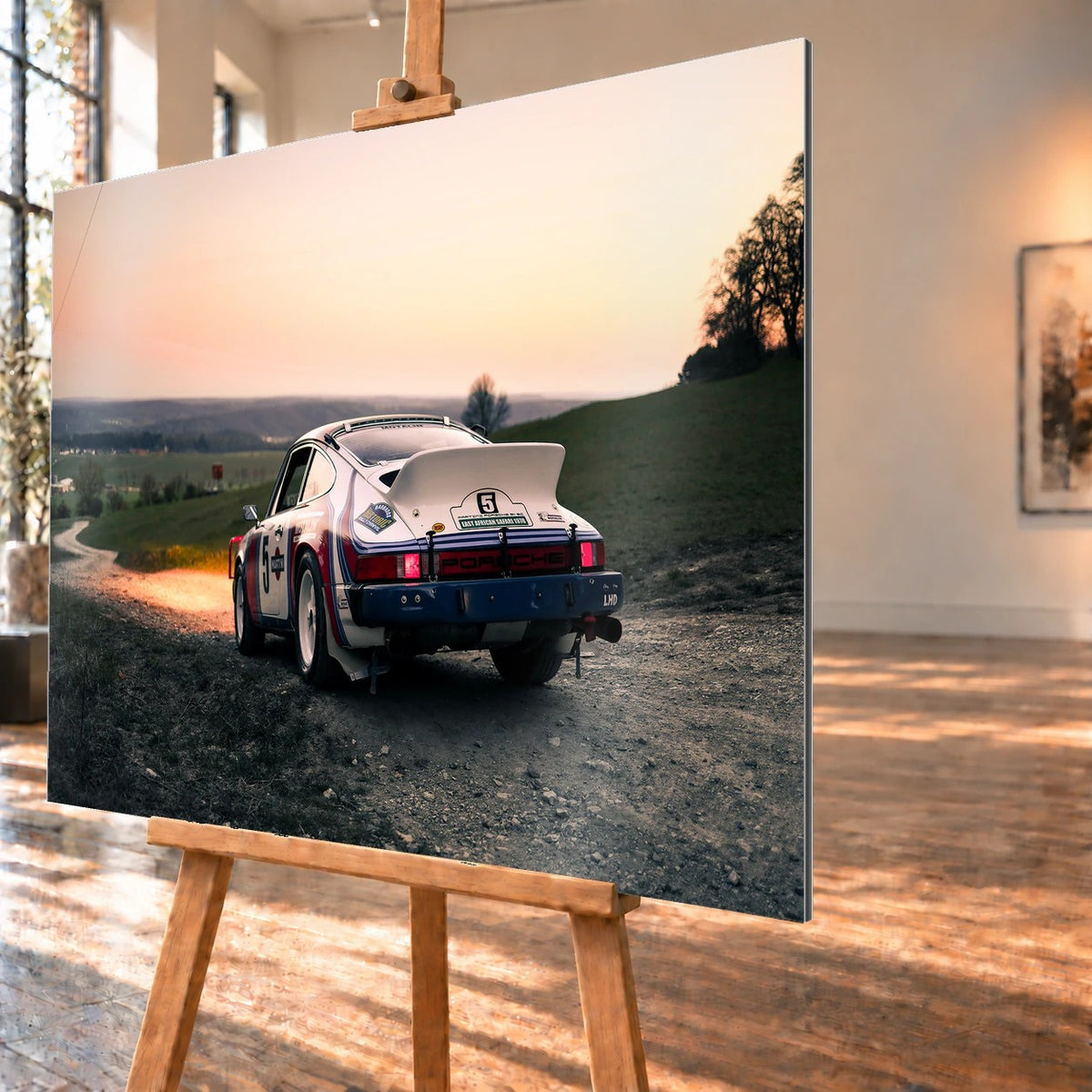 Der Porsche 911 Safari steht von hinten auf einer Anhöhe im Abendrot; weiter Himmel und orangefarbene Horizontlinie öffnen das Bild. Motiv „Fast Motion“ als Alu gebürstet-Wandbild, schräg von der Seite auf einer Holzstaffelei.